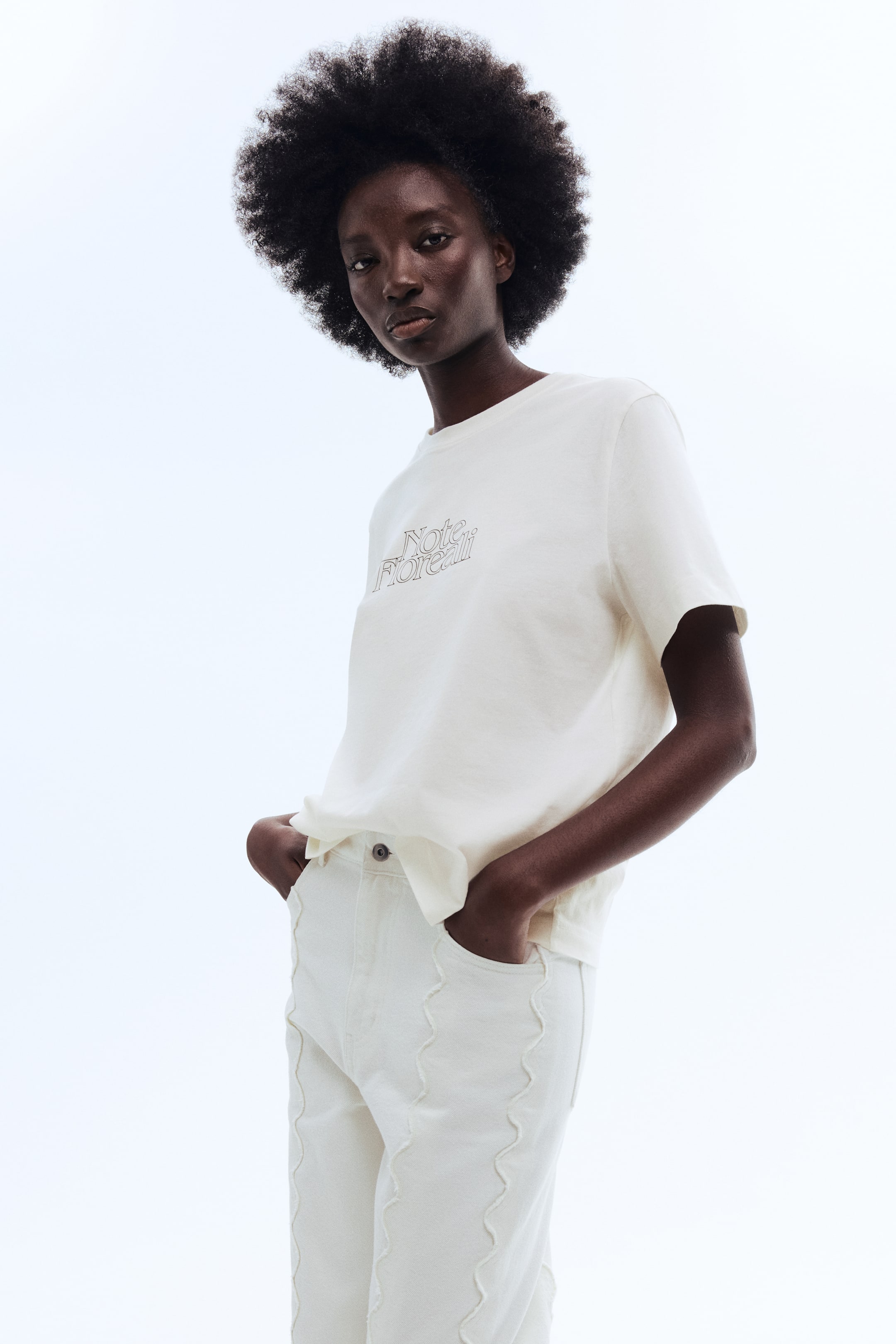 View larger image: A cream-colored t-shirt is showcased from a low angle, featuring a crew neck and short sleeves, with the text 'Note Floreali' in a brown outline font centered on the chest. The t-shirt is styled tucked into pants, highlighting its relaxed fit.