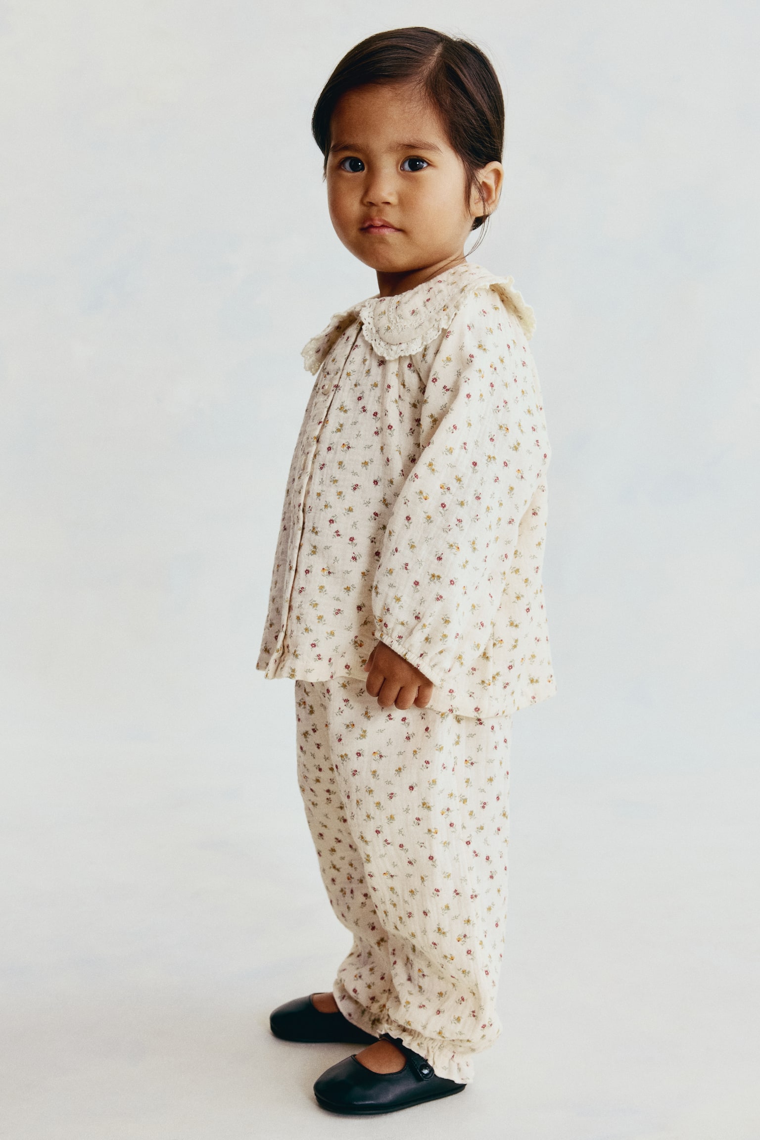 Een bijpassende set van een crÃ¨mekleurige blouse en broek in een katoenweefsel met bloemenpatroon wordt getoond op een kind dat rechtop staat en naar rechts kijkt. De blouse met lange mouwen heeft een afgeronde kraag met kantafwerking en elastische manchetten, terwijl de bijpassende broek elastische pijpuiteinden met rucheafwerking heeft. - 1