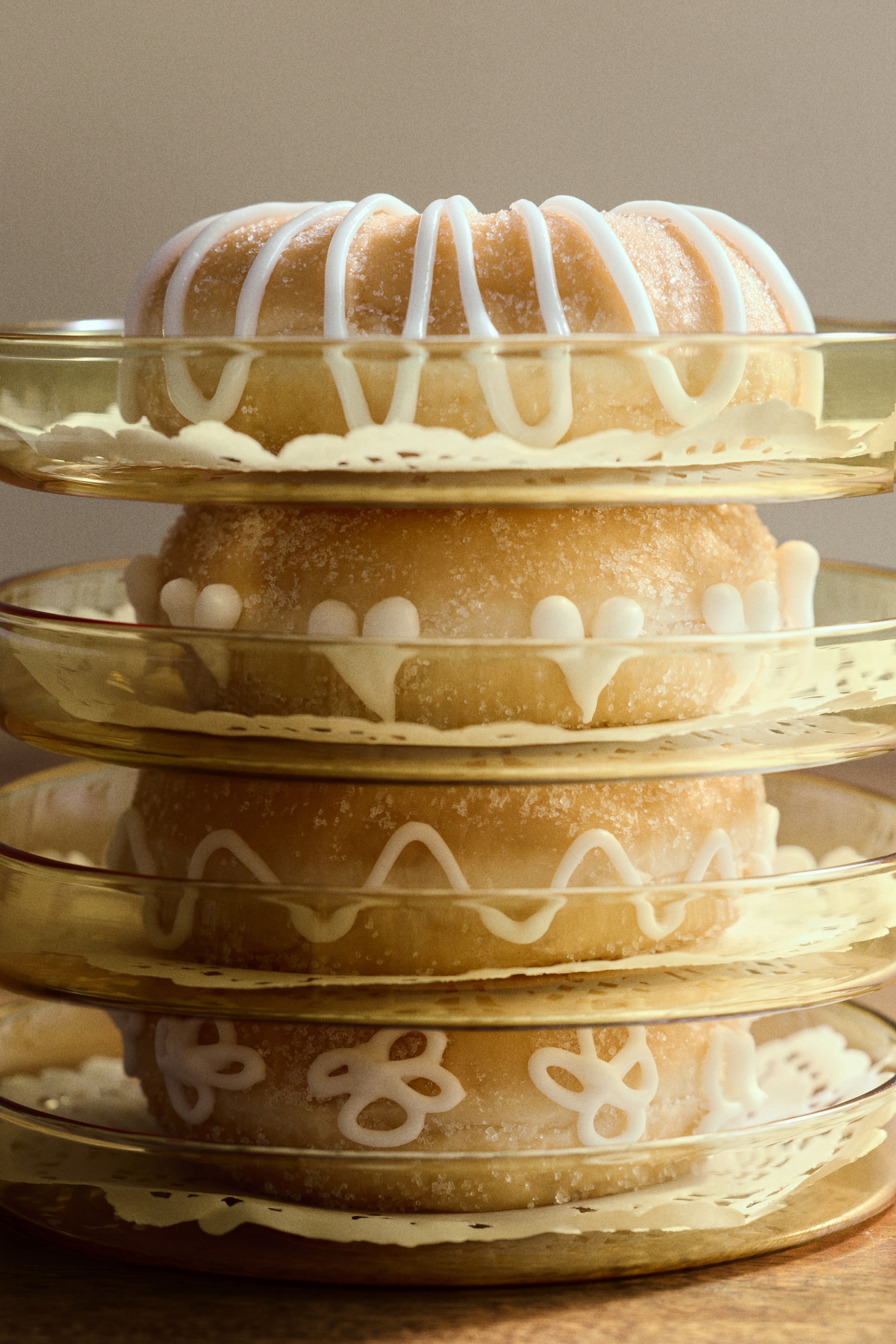 Voir une image plus grande: Une pile de quatre gâteaux bundt dorés et bruns présente des motifs uniques de glaçage blanc et un enrobage de sucre visible. Chaque gâteau repose sur une napperon en papier blanc et une assiette en verre de couleur ambre clair.