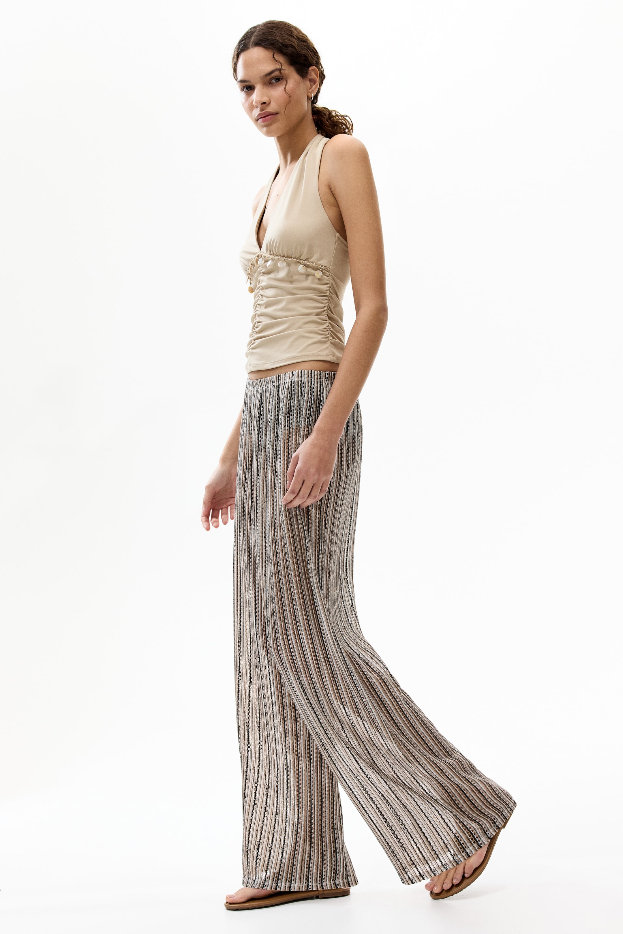 View larger image: A beige halter neck top menampilkan garis leher V, ruched di bahagian depan, dan elemen hiasan kecil di sepanjang tengah. Ia digayakan dengan seluar berpotongan lebar yang berjalur menegak, mempamerkan pelbagai warna coklat, beige, dan hitam dengan tekstur berkilau halus dan mengalir longgar.