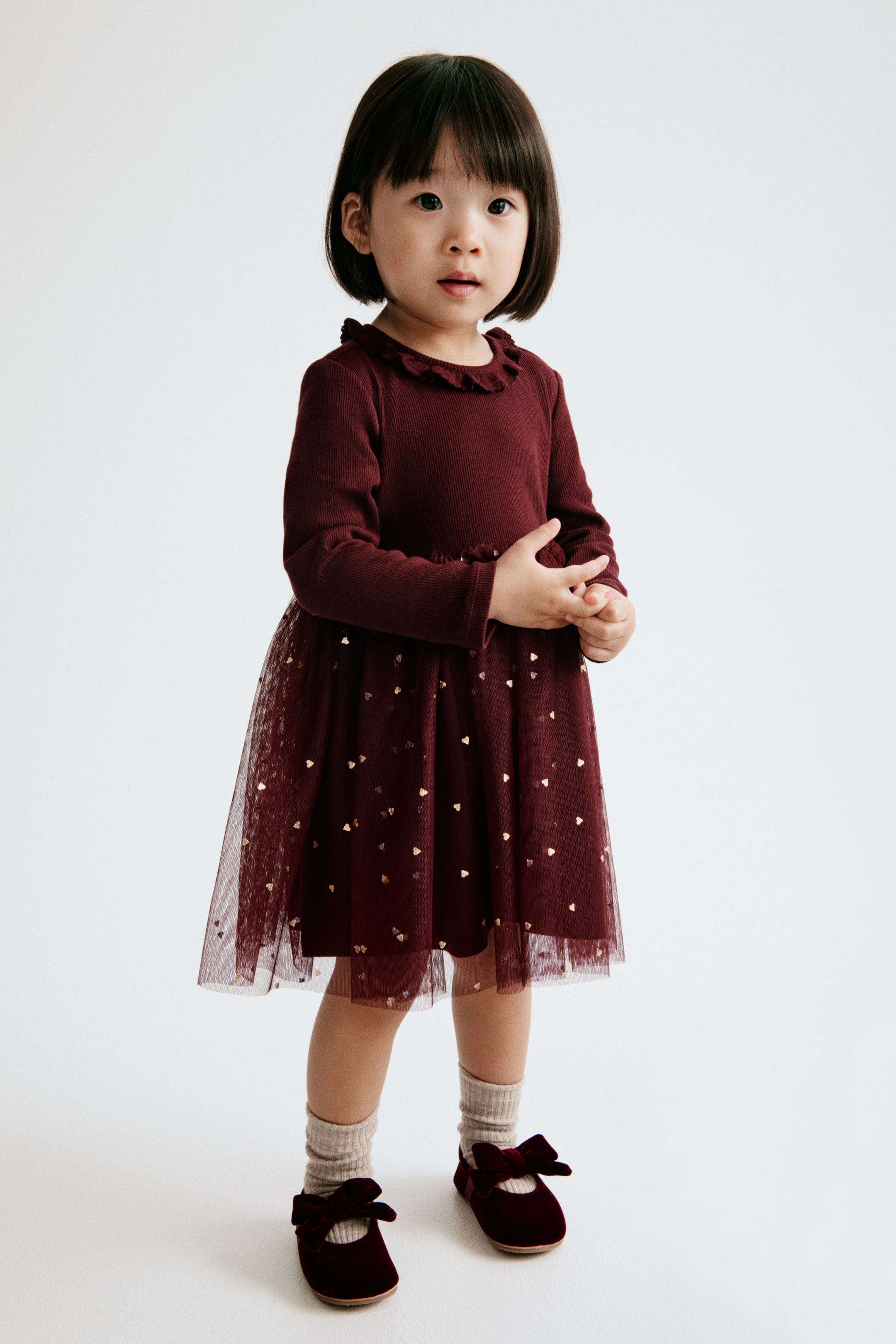 Agrandir l'image: Cette robe bordeaux à manches longues, vue de face, présente un corsage en jersey côtelé avec un col rond bordé de volants et une taille assortie également agrémentée de volants. La robe se prolonge par une jupe en tulle évasée et doublée, décorée de petits cœurs dorés scintillants.