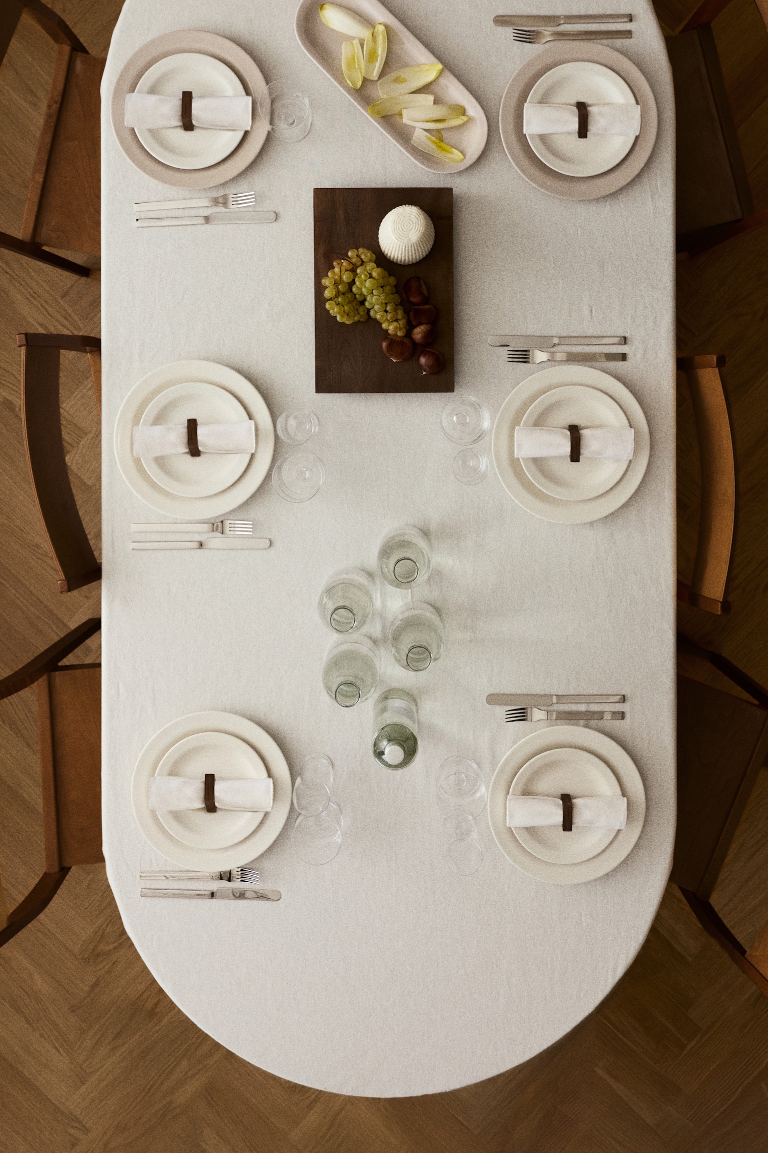An off-white, textured linen tablecloth is draped over an oval dining table, presenting six symmetrical place settings. Each setting comprises stacked ceramic plates, a rolled white napkin secured with a dark brown ring, and silver-toned cutlery, with glass bottles and serving dishes arranged along the centre. - 1