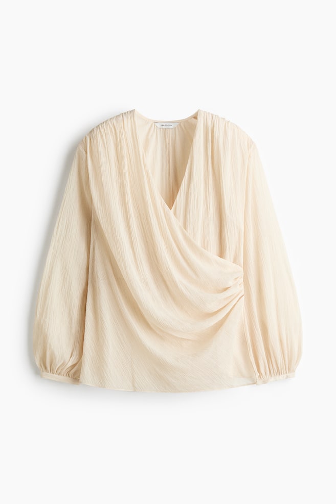 A cream-coloured blouse in a crinkled weave is shown from the front, featuring a deep V-neckline with a sewn-in wrap front that creates a draped effect with gathered side seams. The long balloon sleeves taper into narrow cuffs at the wrists. - 1