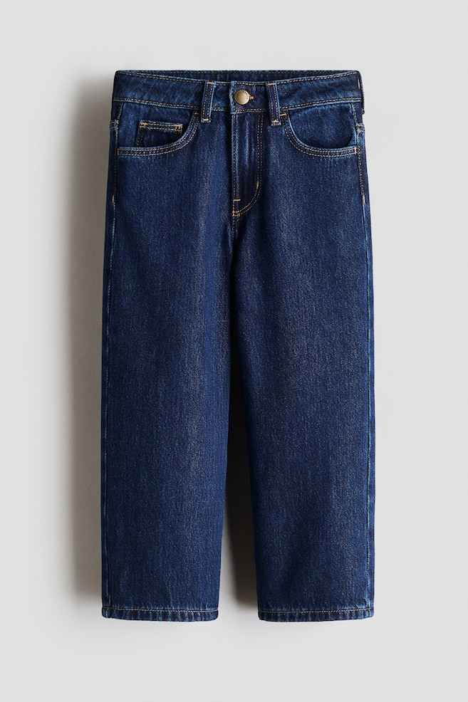 Jean en denim bleu foncé posé à plat, offrant une vue de face. Il présente un bouton doré, des passants de ceinture, deux poches avant et deux petites poches à monnaie, avec des surpiqûres contrastantes couleur tan soulignant ces détails. - 1