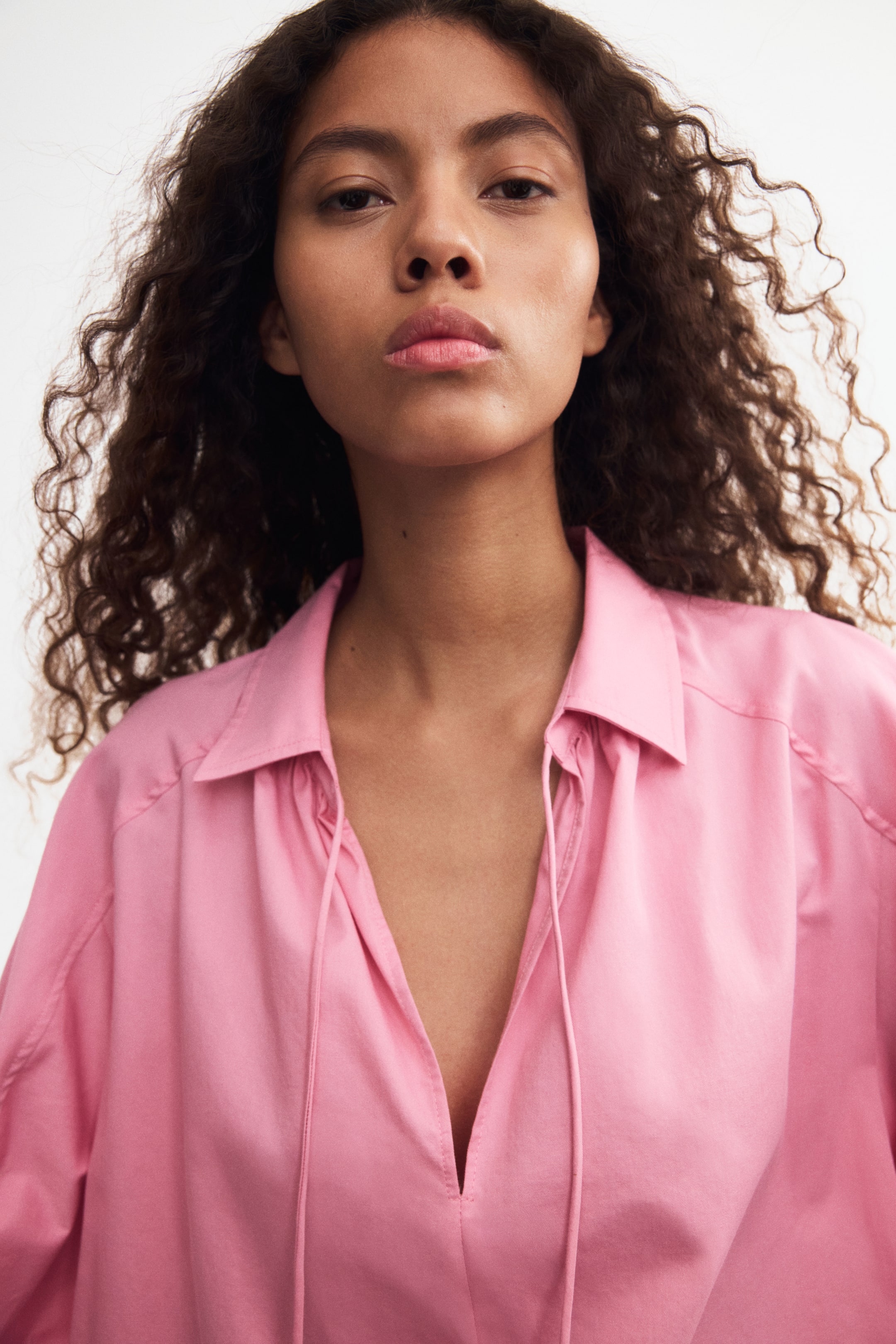Visualizza immagine più grande: La modella indossa una blusa rosa con colletto, caratterizzata da uno scollo a V con due lacci regolabili visibili che pendono morbidamente.
