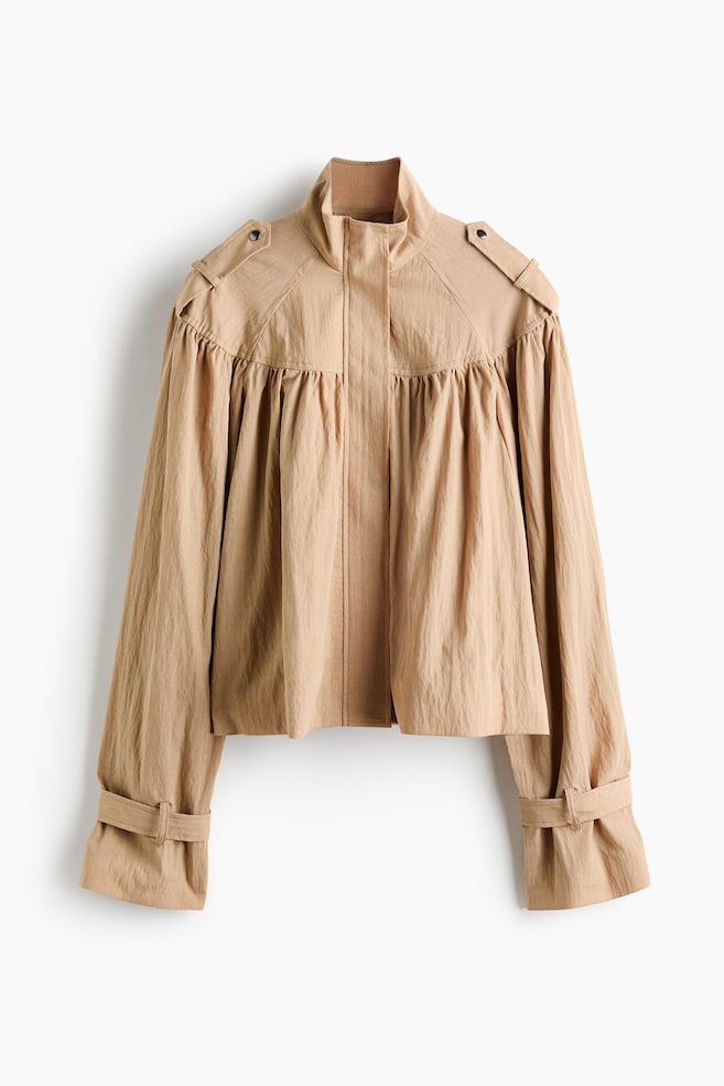 A beige cropped jacket is hanging facing front, featuring a high stand-up collar, shoulder epaulets, and a concealed front placket. The design showcases prominent gathering below the yoke that creates a voluminous drape throughout the body and wide sleeves, which are finished with adjustable cuff straps. - 1