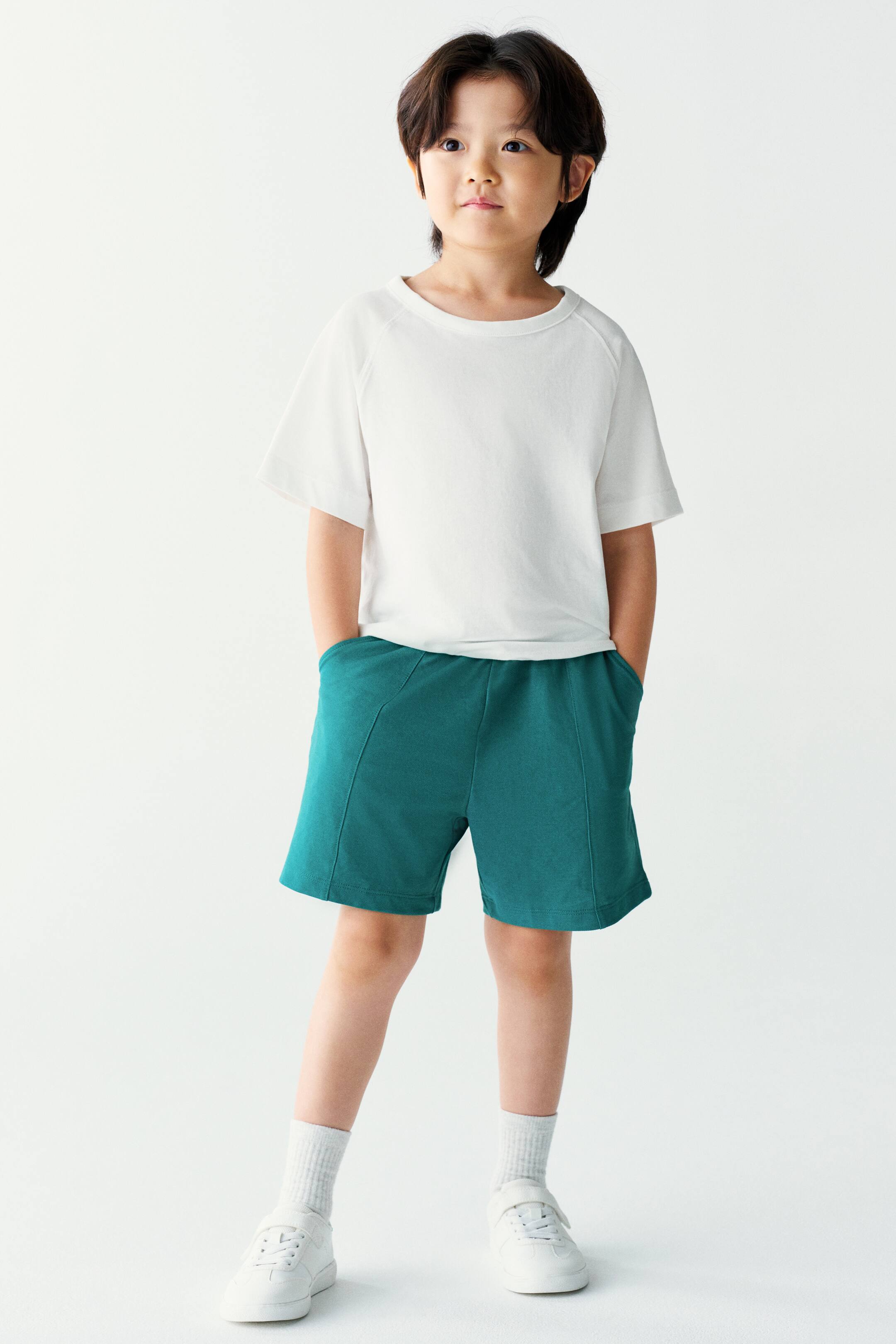 View larger image: The model is wearing a white short-sleeved T-shirt and teal green knit shorts, standing with hands resting in the side pockets. The casual look is styled with white crew socks and white sneakers.