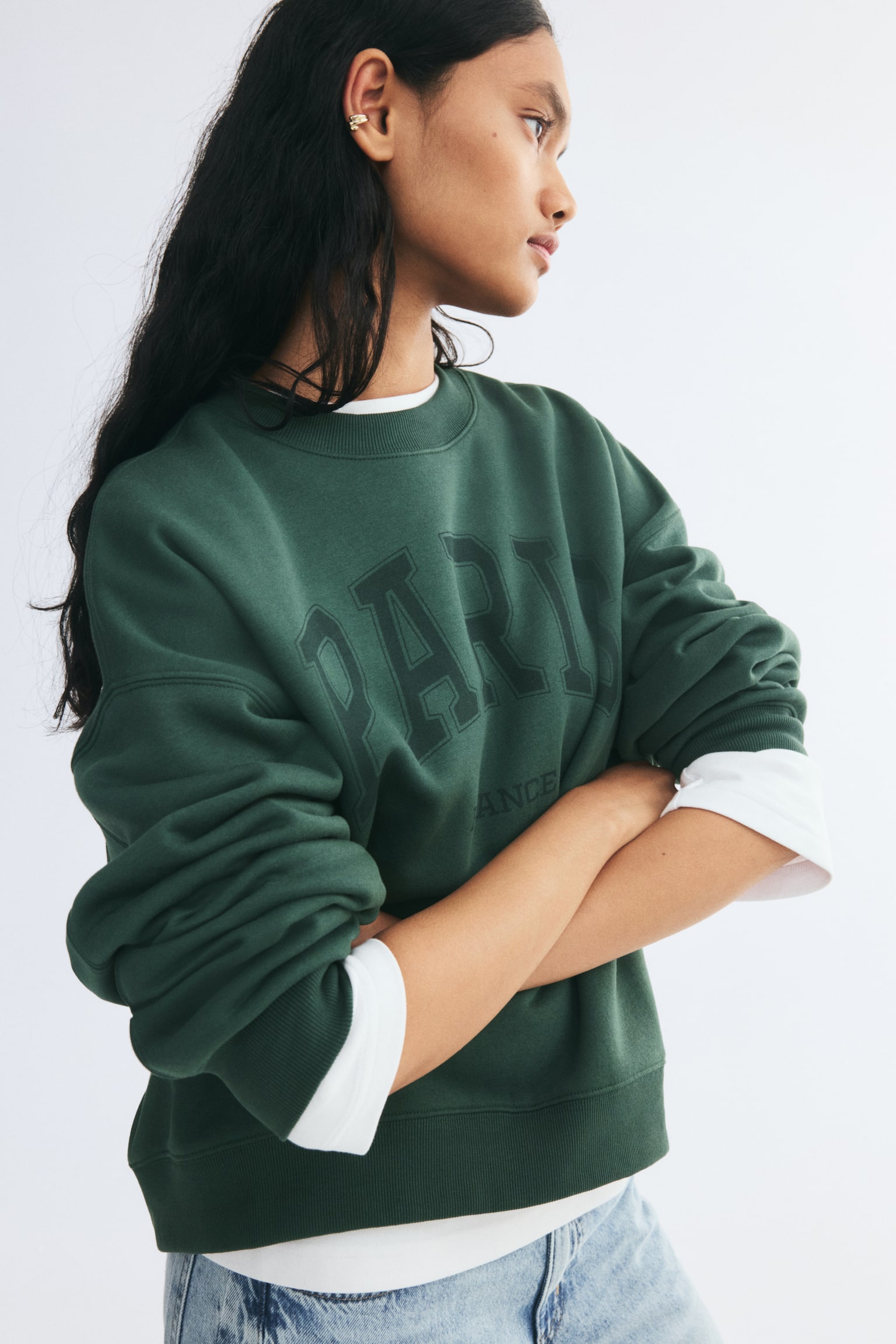 A dark green oversized sweatshirt with a printed "PARIS" graphic on the front is worn by the model with folded arms. The sweatshirt features dropped shoulders, a ribbed neckline, cuffs, and hem, and is styled over a white long-sleeve top paired with light-wash jeans. - 1