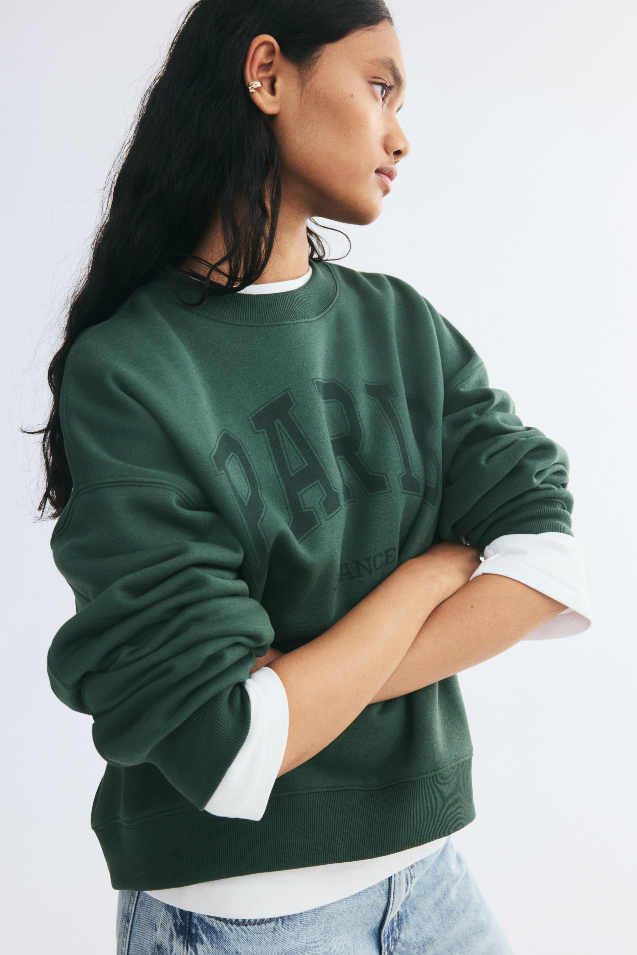 View larger image: A dark green oversized sweatshirt featuring a printed "PARIS" graphic on the front is worn by the model with folded arms. The sweatshirt has dropped shoulders, a ribbed neckline, cuffs, and hem, and is styled over a white long-sleeve top and paired with light-wash jeans.