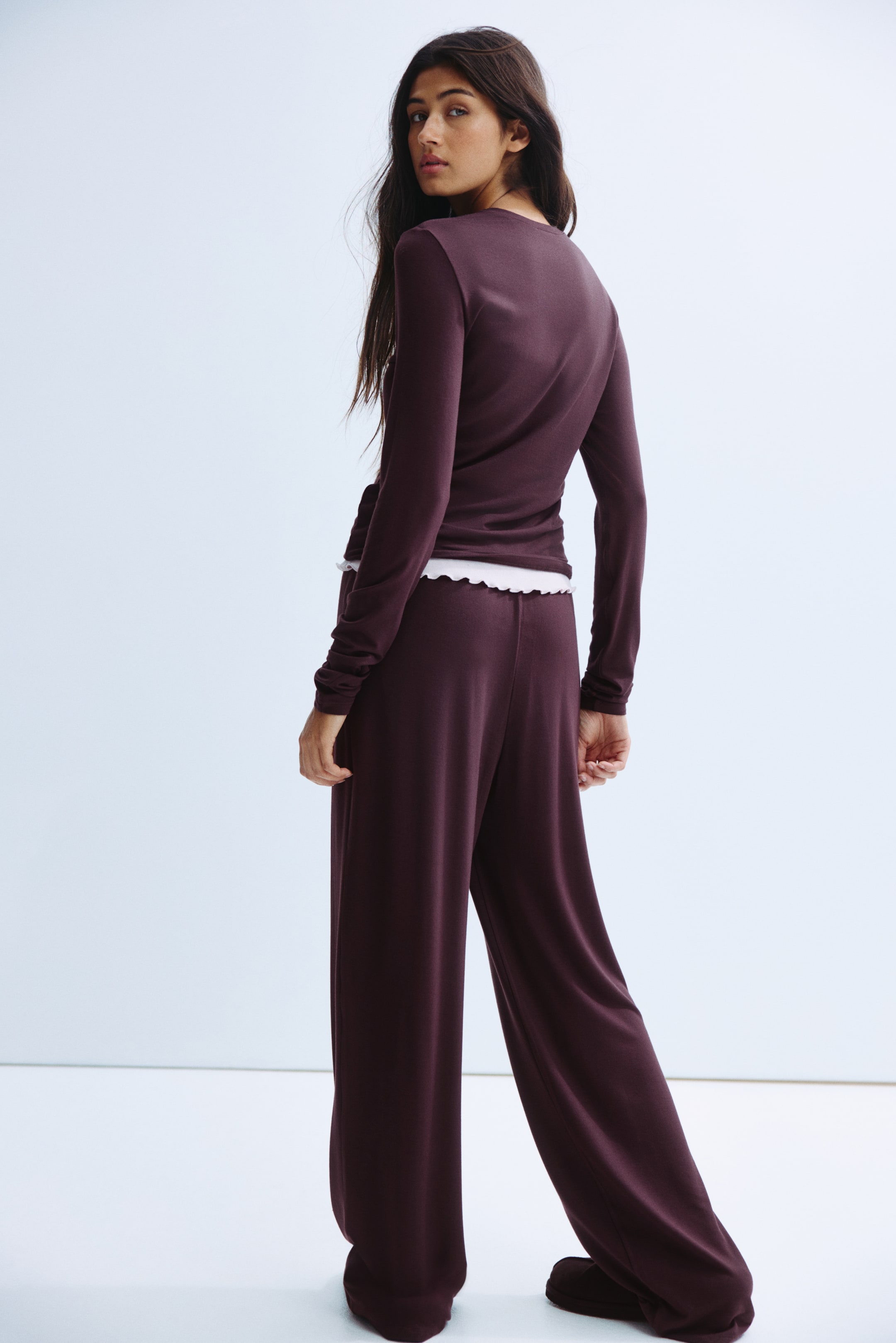 Grotere afbeelding bekijken: Een donker pruimen tweedelig loungewear set wordt gepresenteerd vanuit een driekwart achterhoek. Het heeft een lange mouwen top met een zijstrik, die een witte gerimpelde zoom eronder onthult, en bijpassende wijde pijpen broek.