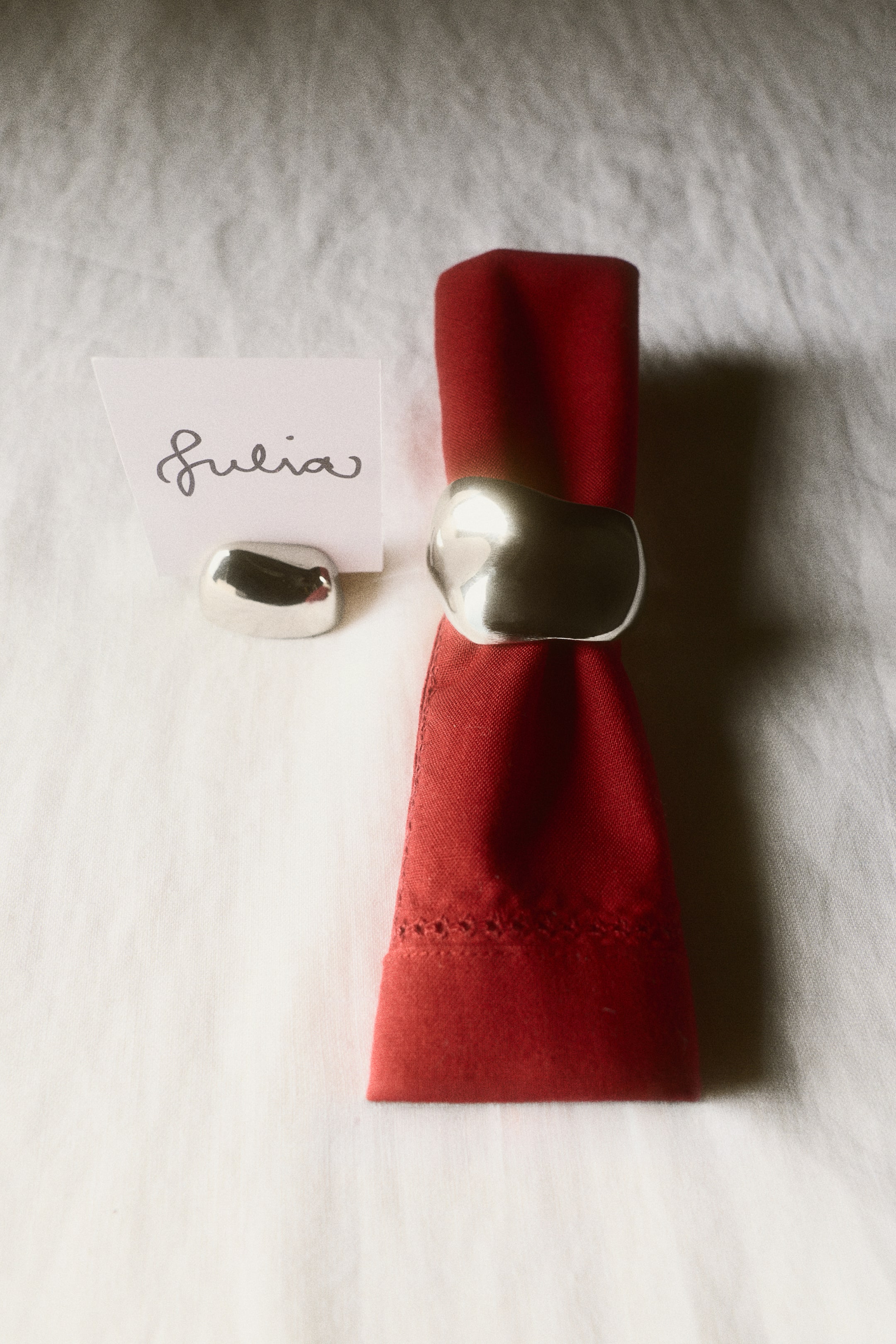 View larger image: A polished stainless steel dining accessory set featuring an organic, sculptural design, with a napkin ring holding a folded red fabric napkin and a matching place card holder holding a small white card with handwritten text.