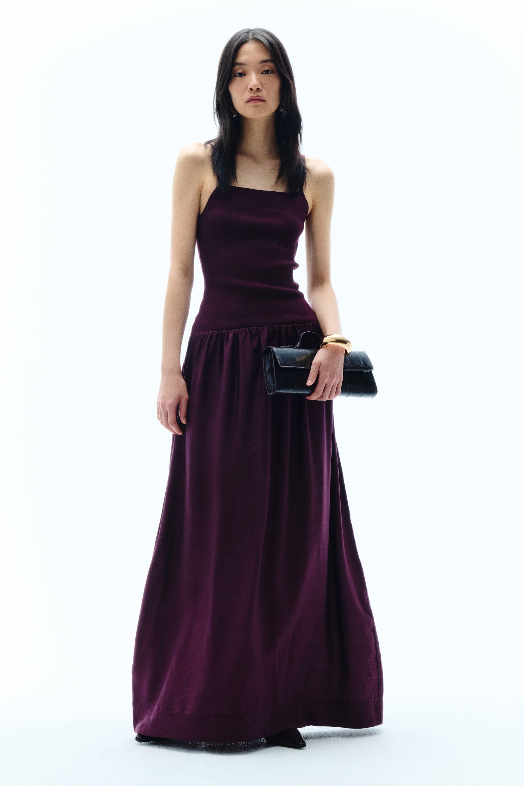 View larger image: A deep purple, full-length dress is shown from the front, featuring a form-fitting, ribbed knit bodice with thin shoulder straps and a straight neckline. The bodice cinches at the waist, flowing into a gathered skirt that extends to the floor in a classic A-line silhouette.