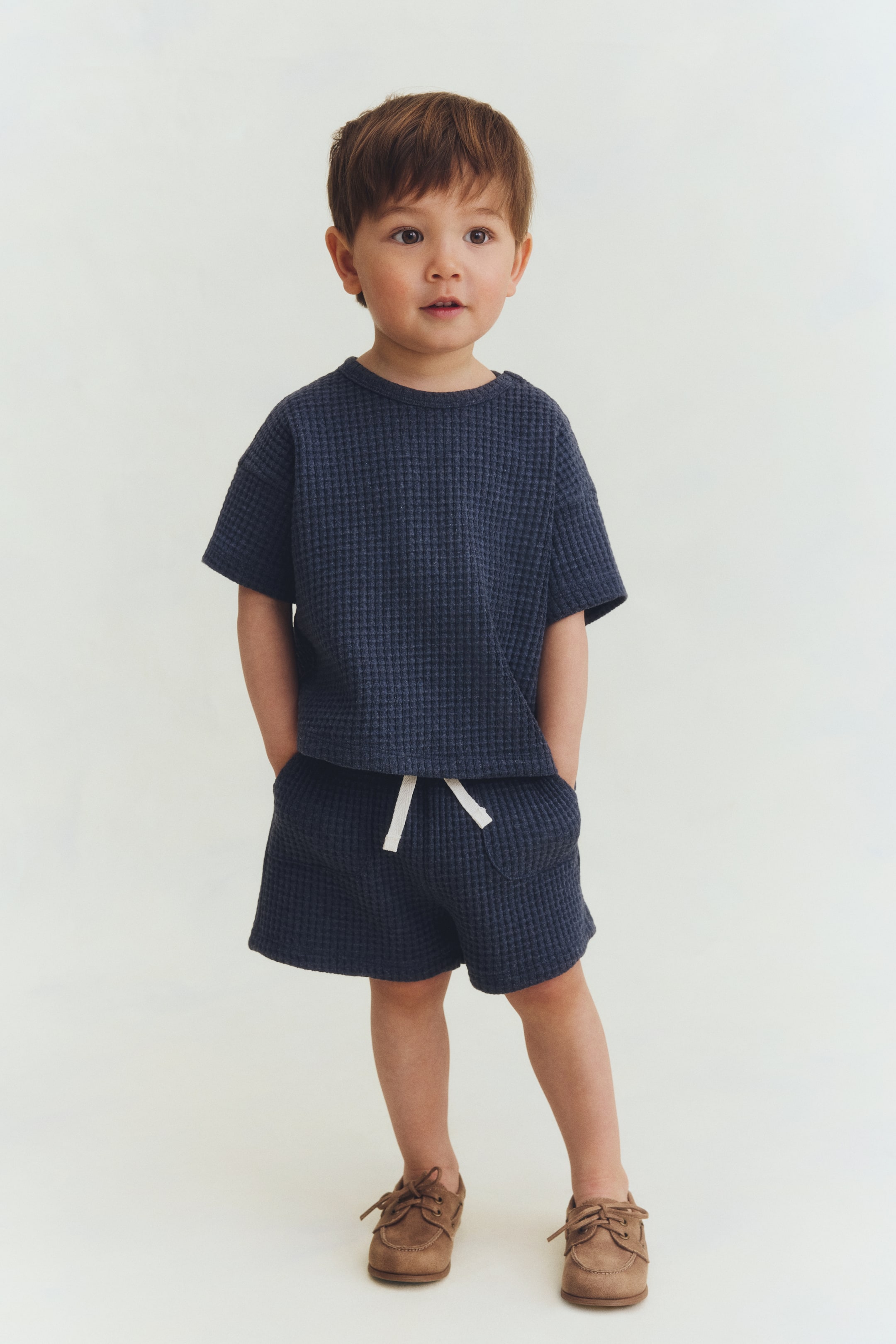 Grösseres Bild ansehen: Ein dunkelblaues Waffelstrick-Set mit kurzärmligem T-Shirt und passenden Shorts präsentiert sich in einer entspannten Passform. Das T-Shirt verfügt über einen Rundhalsausschnitt, und die Shorts sind mit einem elastischen Bund, einem weißen Kordelzug und Fronttaschen ausgestattet.