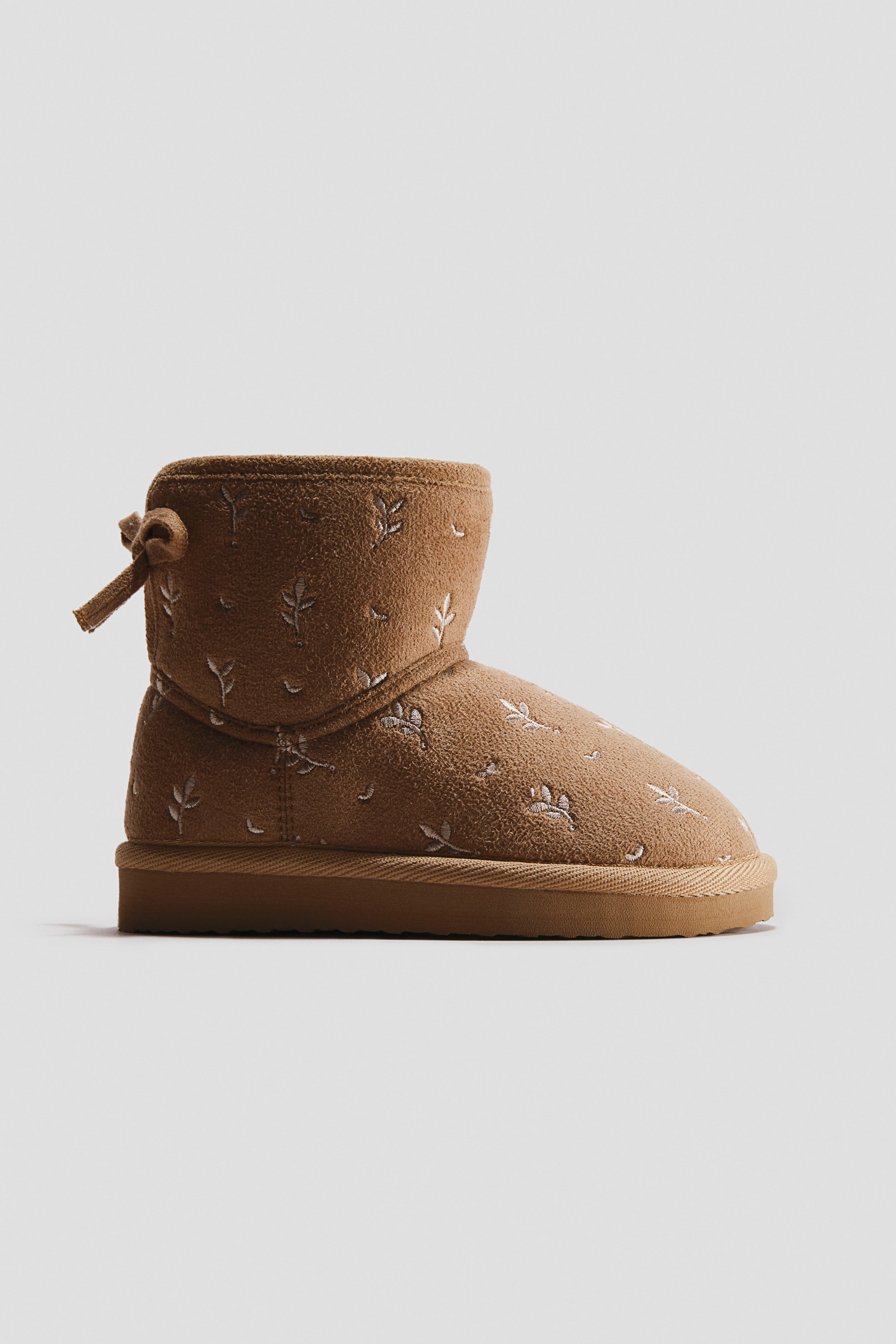 View larger image: An ankle-high boot with a brown napped finish is shown in side profile facing right, featuring small white embroidered leaf patterns and a decorative fabric bow at the back collar. The boot is constructed with a thick, textured tan rubber sole.