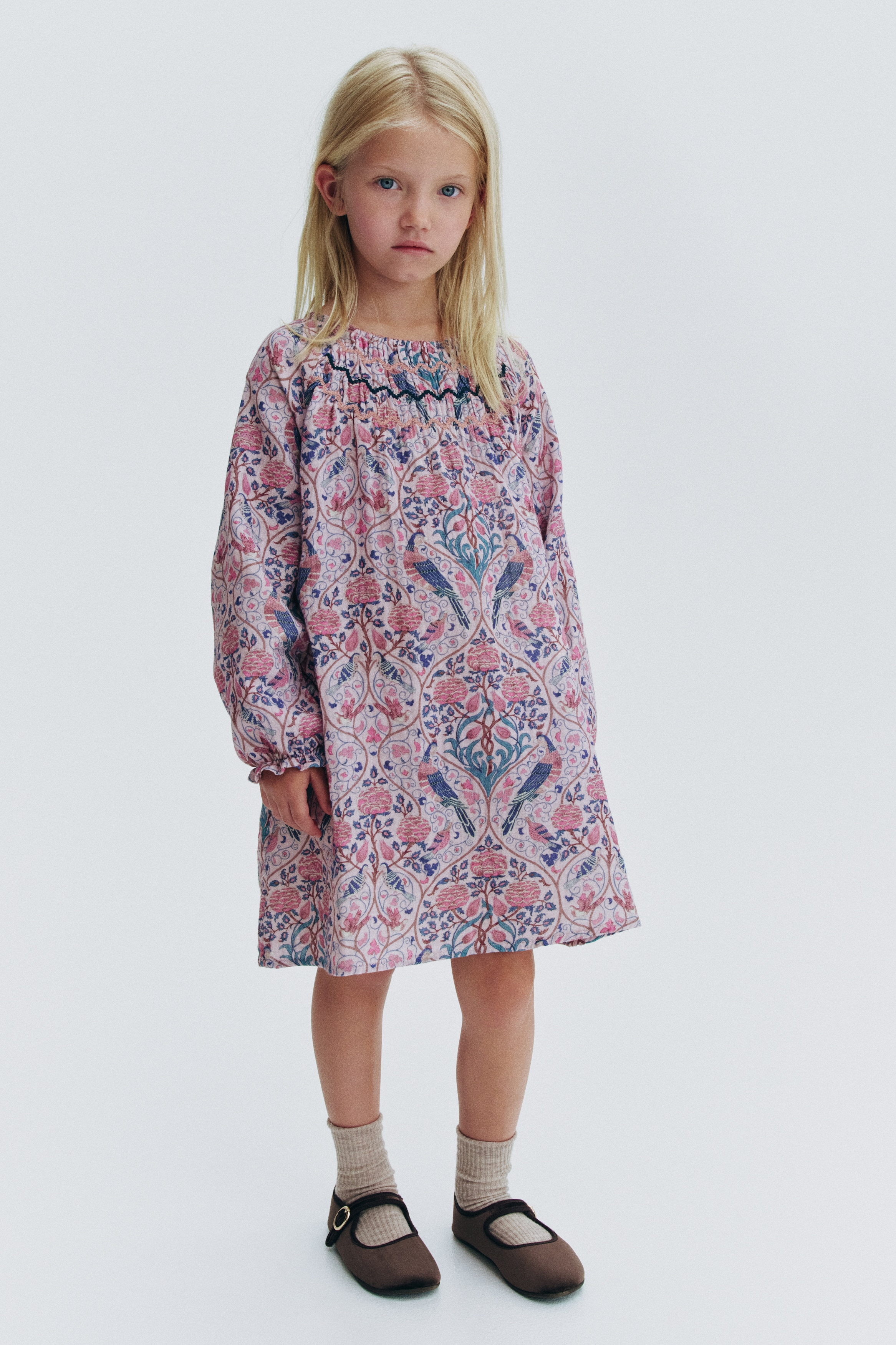An A-line dress showcases an all-over floral and bird print in shades of pink, teal, and blue, shown from the front. The dress features a round neckline with picot trim, a smocked chest panel with embroidered detailing, and long raglan sleeves with elasticated, frill-trimmed cuffs.