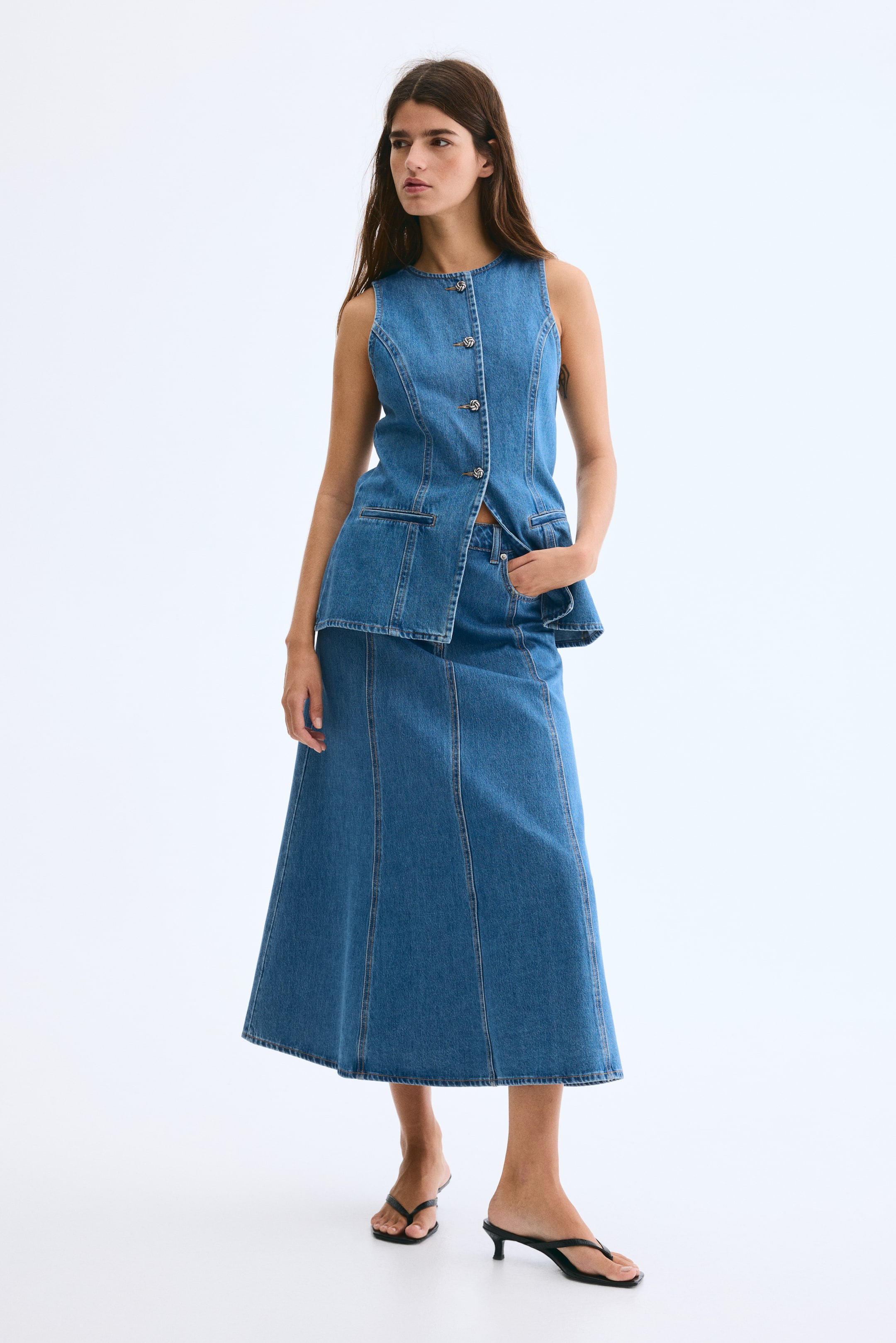 View larger image: A coordinated medium-wash denim set, comprising a sleeveless vest and an A-line midi skirt, is being presented from a front-facing angle. The vest is featuring a round neckline, a front button placket with five decorative buttons, and two flap pockets, with its hem flaring out from the waist. The midi skirt is designed with vertical panel seams, extending to a midi length.
