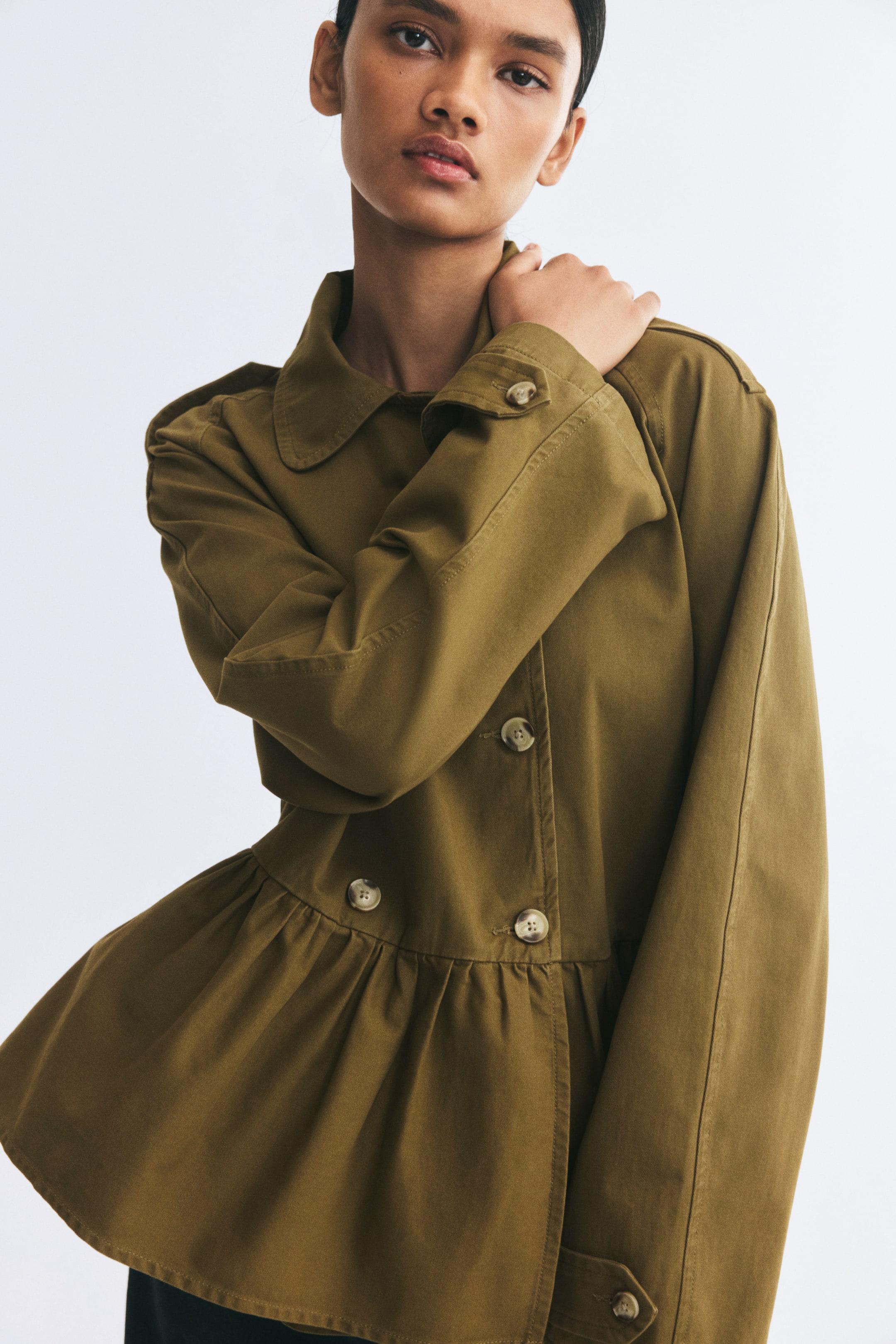 View larger image: The model is wearing a short, double-breasted trench coat in olive green, with one hand resting on their shoulder. The coat features a collar, buttoned shoulder tabs, a double-breasted front button closure, and a gathered seam at the waist with a flared peplum.