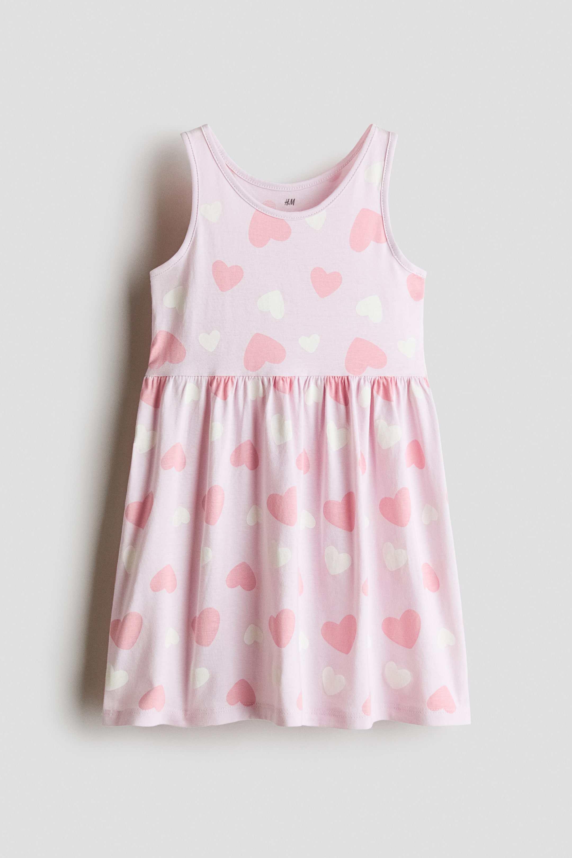 View larger image: A light pink sleeveless jersey dress is shown from the front, featuring an all-over pattern of scattered pink and white hearts. The dress is designed with a round neckline, wide shoulder straps, and a gathered seam at the waist that transitions into a flared skirt.