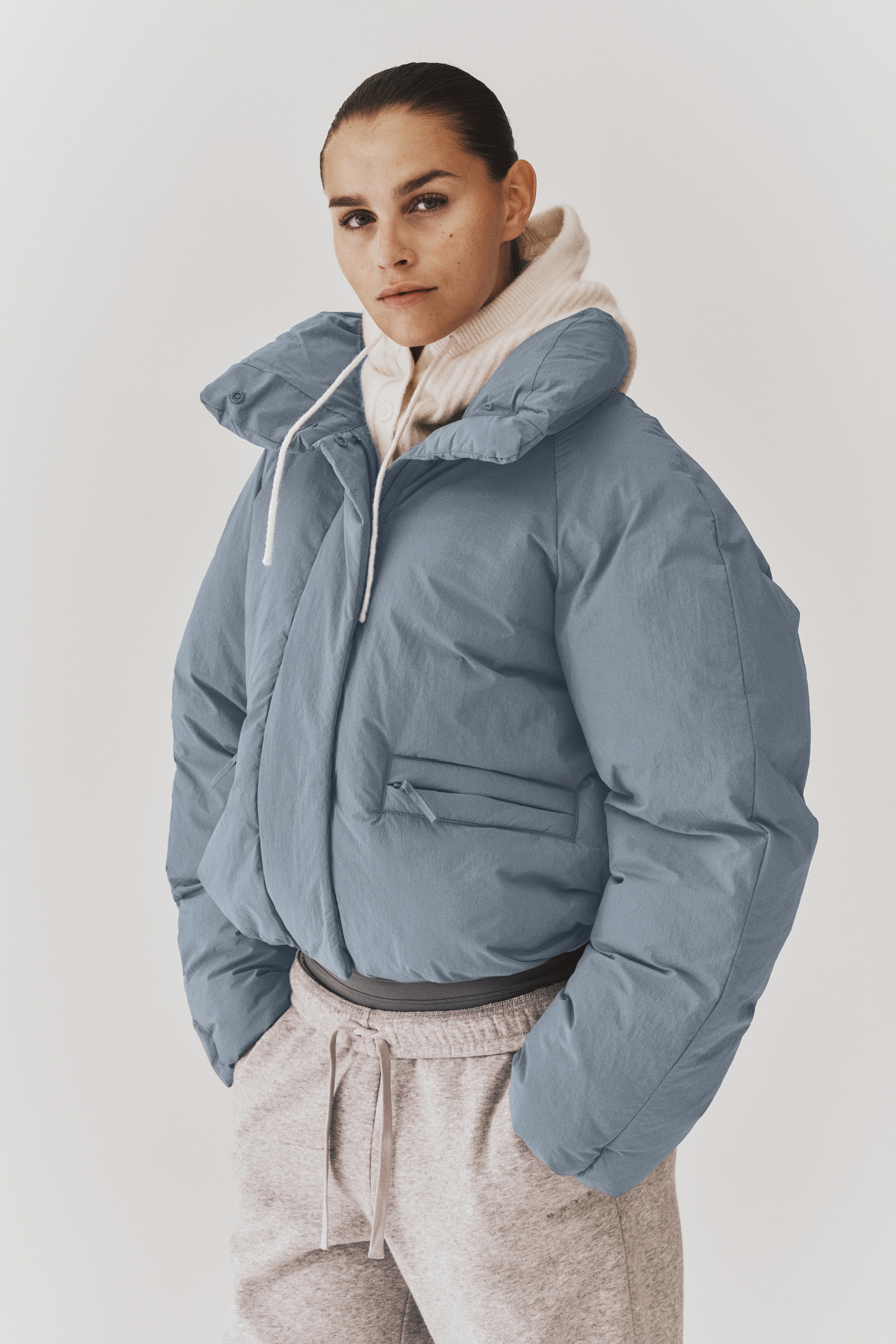 The front-facing cropped puffer jacket features a high, padded stand collar, a hidden front zipper closure, and raglan sleeves. It displays a matte slate-grey finish and two horizontal zippered welt pockets on the lower front.