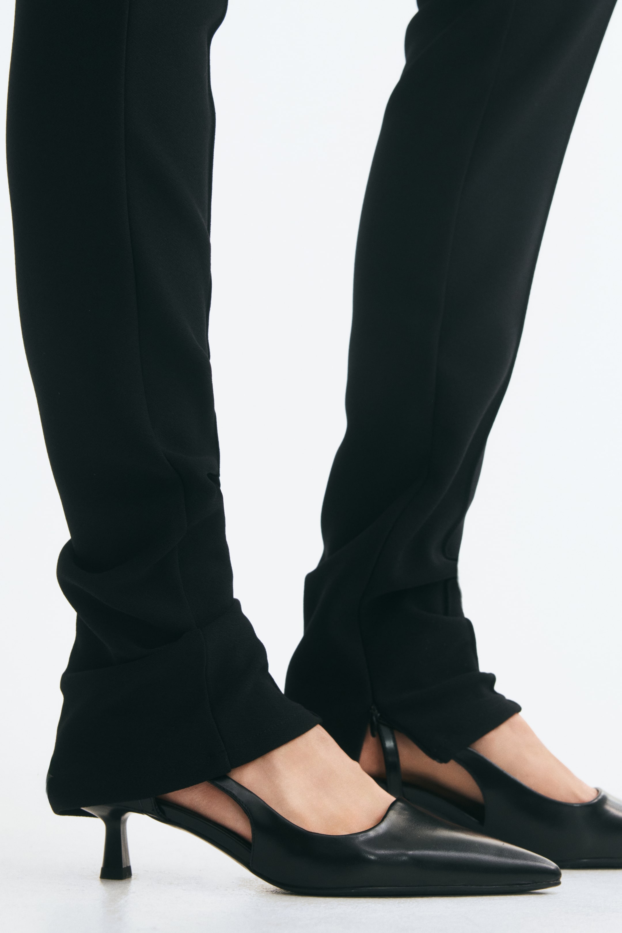 Vis større bilde: Svarte slingback-sko i belagt stoff bæres av modellen, med spisse tåpartier, smale hælremmer og lave kitten-heels. Skoene styles med smale svarte bukser som faller over baksiden av anklene.