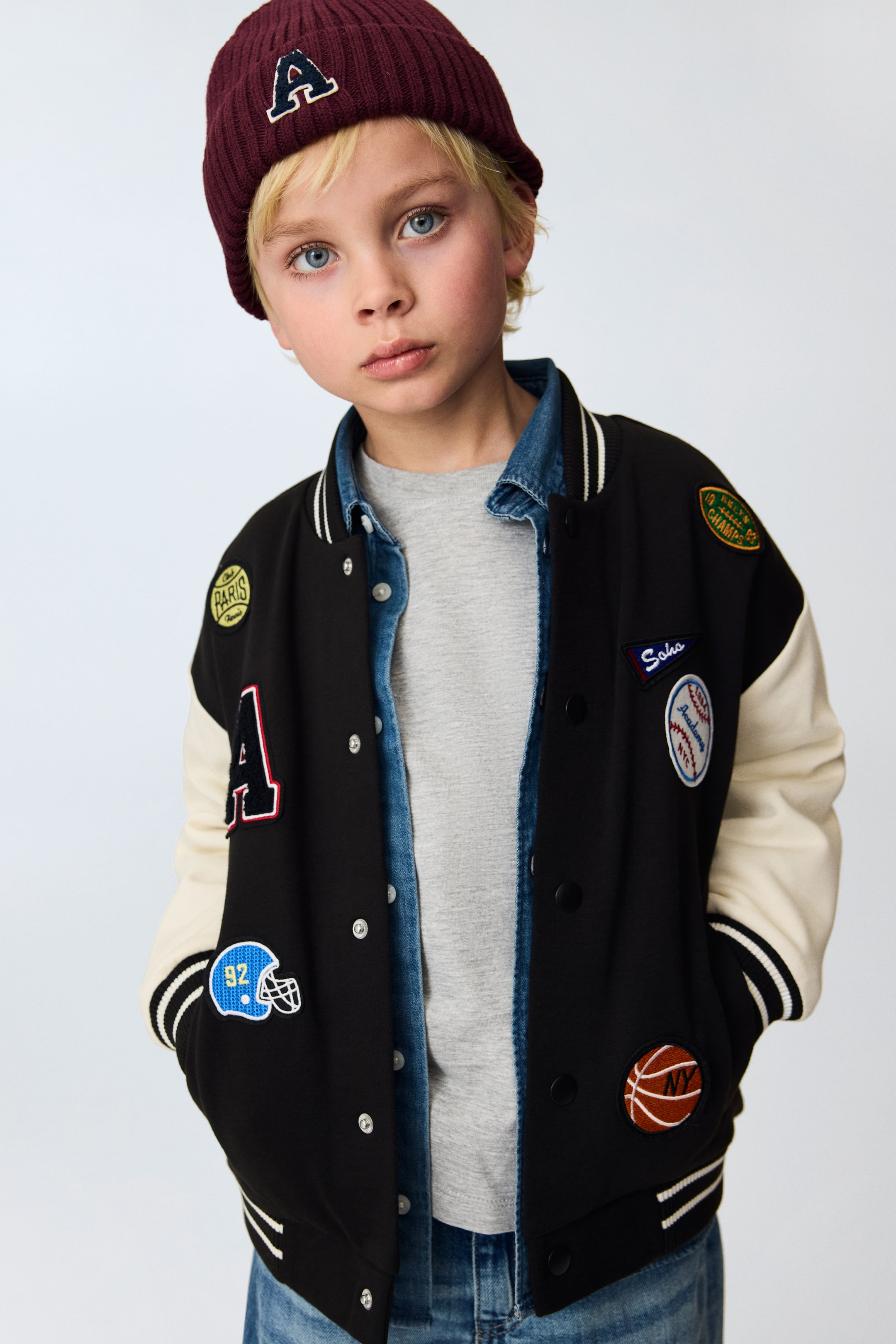 View larger image: A black varsity jacket with cream sleeves features a snap button front closure and white double stripes on its ribbed cuffs and hem. It boasts various patches, including a large red "A" and multiple sports-themed designs, and is styled open over a denim shirt and grey t-shirt.