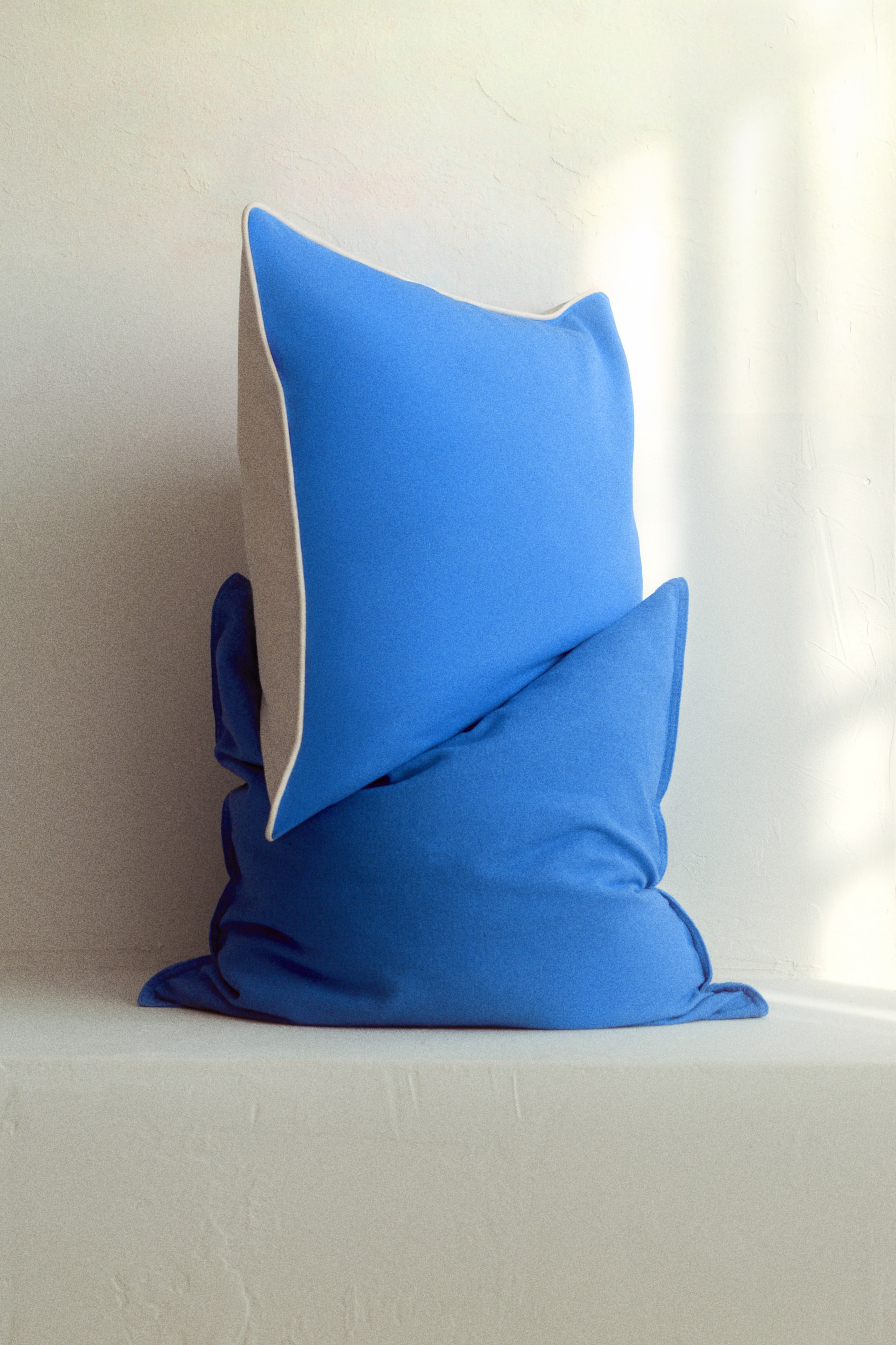 View larger image: A vibrant blue square cushion, featuring a light grey side panel and white piping, is resting on a second, unstructured darker blue cushion. The top cushion is angled slightly to the left.