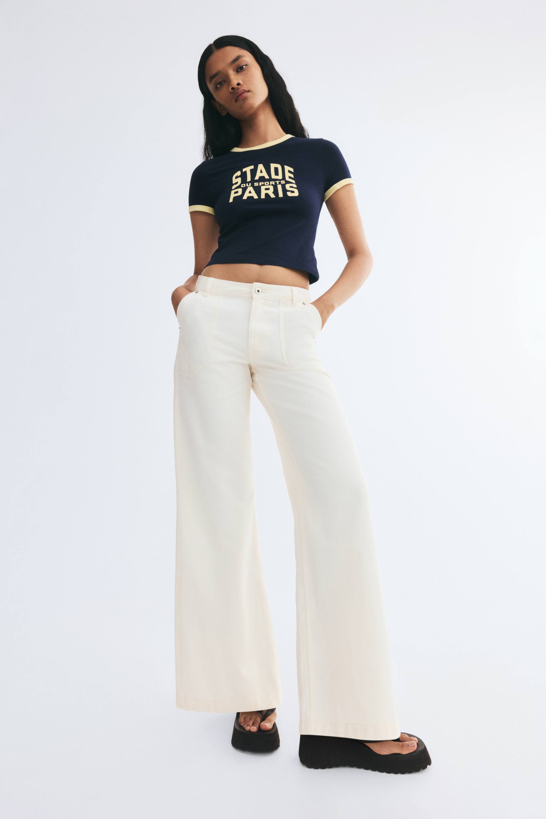 View larger image: Off-white cotton twill pants featuring a high waist, button closure, wide legs, and diagonal side pockets are worn by the model, who is standing with her hands tucked into the pockets. The pants are styled with a dark cropped graphic T-shirt and black platform sandals.