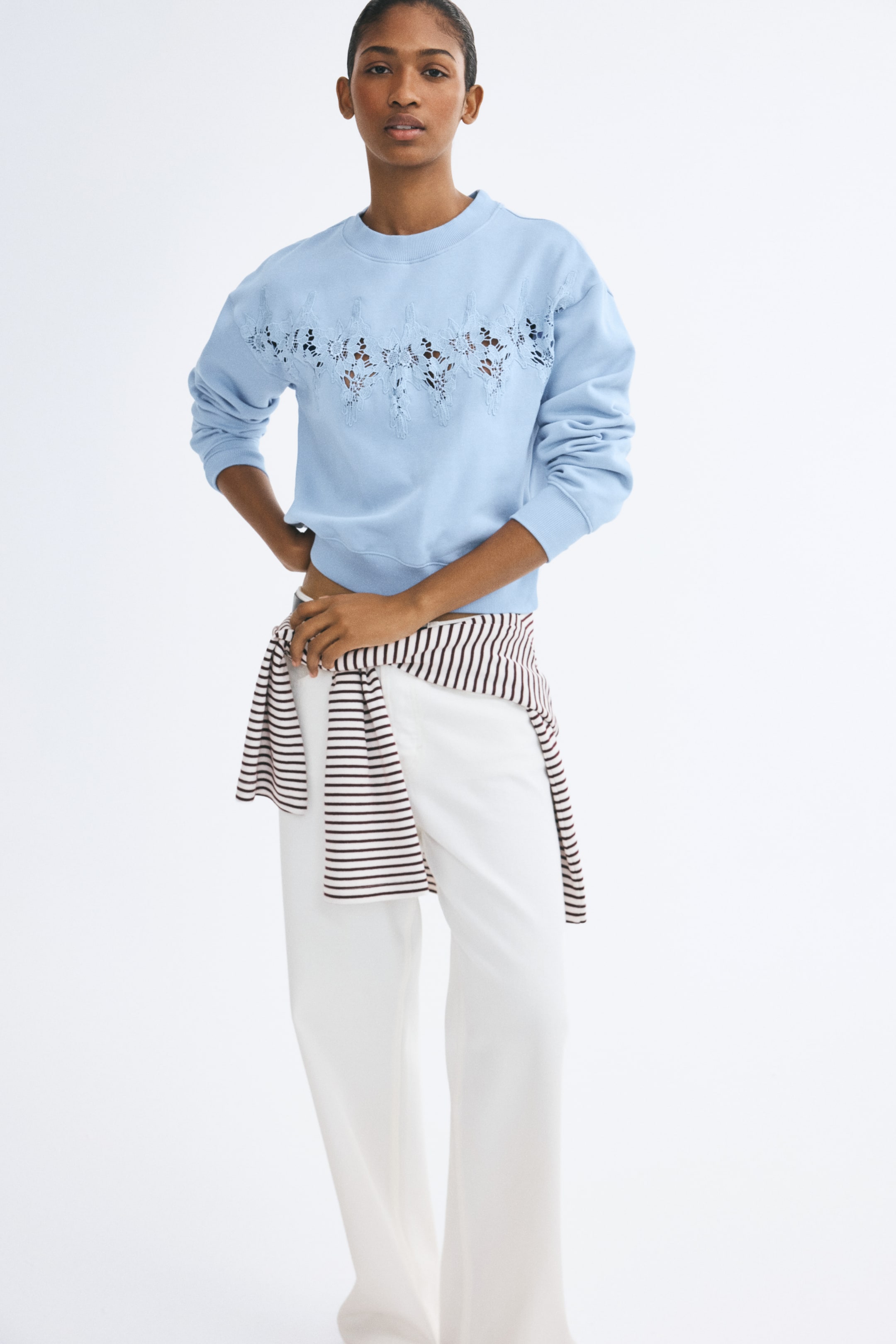 View larger image: A light blue, loose-fit sweater featuring an irregular lace inset across the chest is being worn by the model. The sweater, which has a round ribbed neckline, dropped shoulders, and ribbed cuffs, is styled with white trousers and a striped shirt tied around the waist.