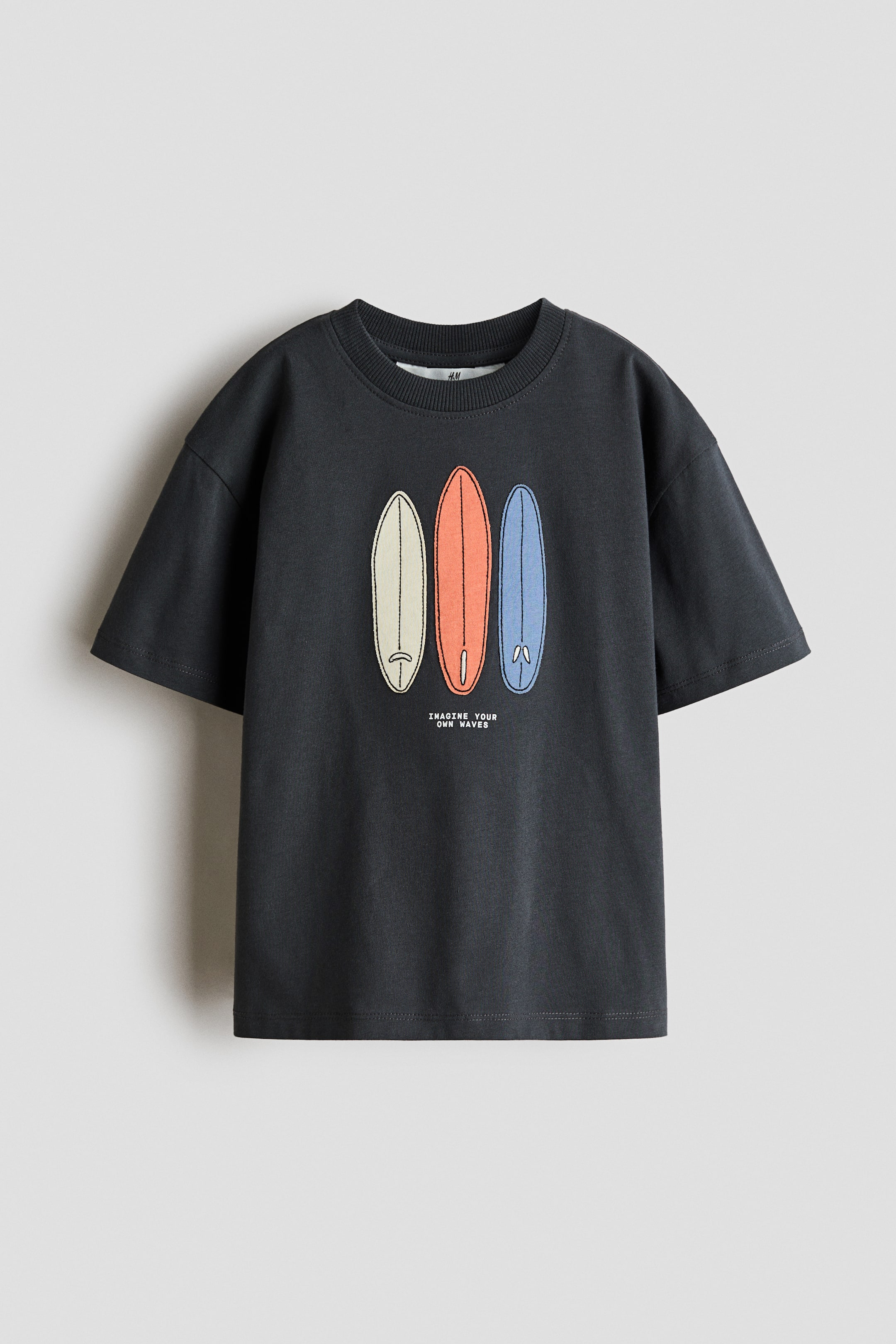 Visualizza l'immagine ingrandita: Una T-shirt in jersey di cotone grigio scuro, a vestibilità ampia, è esposta dal davanti, mostrando uno scollo rotondo con bordo a costine. Il davanti è decorato con un motivo ricamato di tre tavole da surf nei colori crema, corallo e azzurro chiaro, e il testo "IMAGINE YOUR OWN WAVES" è posizionato sotto di esse.