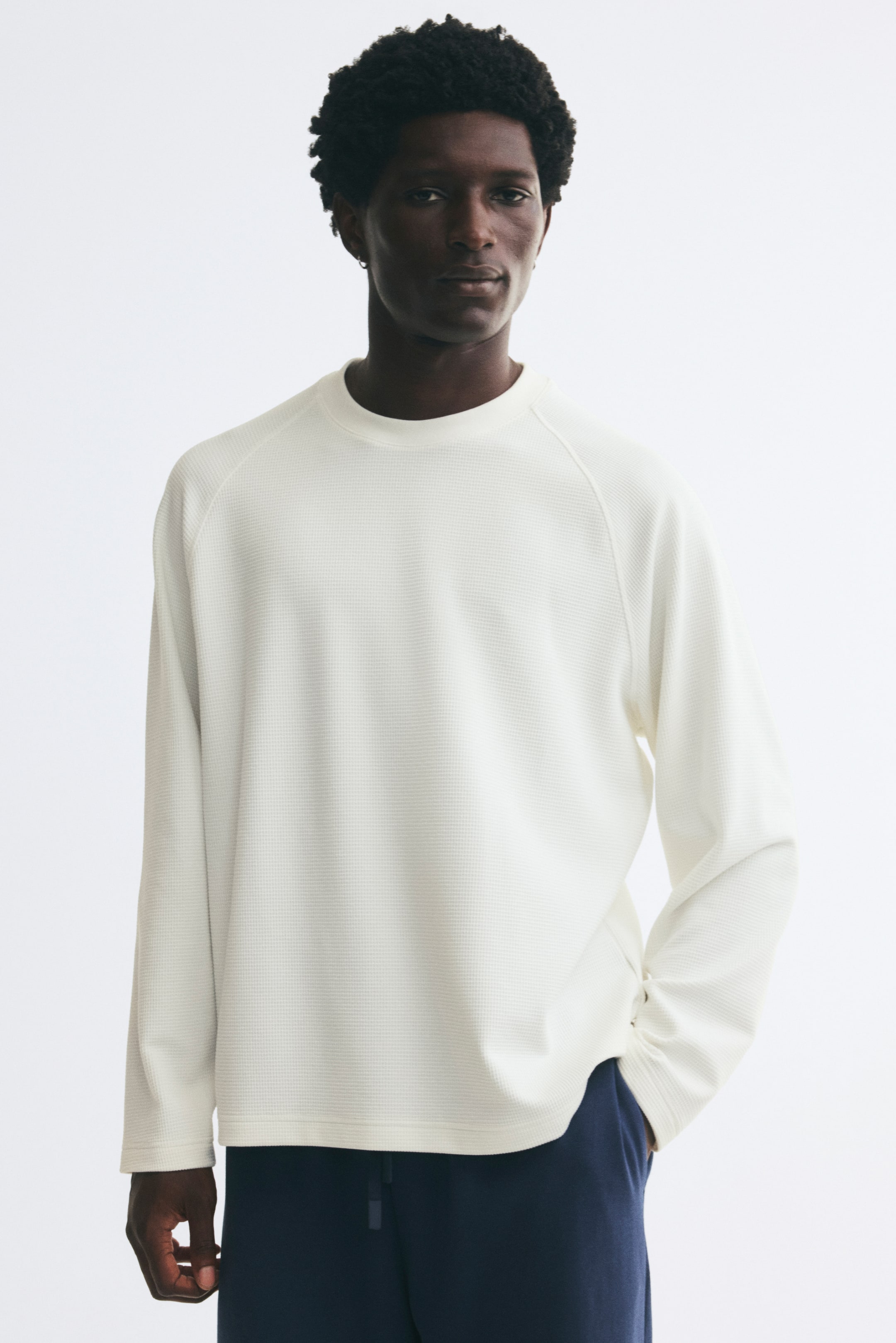 Voir une image plus grande: Un tee-shirt en jersey gaufré blanc cassé, doté d’un col rond, de manches raglan longues et d’un ourlet droit, est porté par le modèle. Le modèle se tient debout, une main glissée dans la poche d’un pantalon bleu foncé, mettant en valeur la coupe décontractée et la silhouette casual de la chemise.