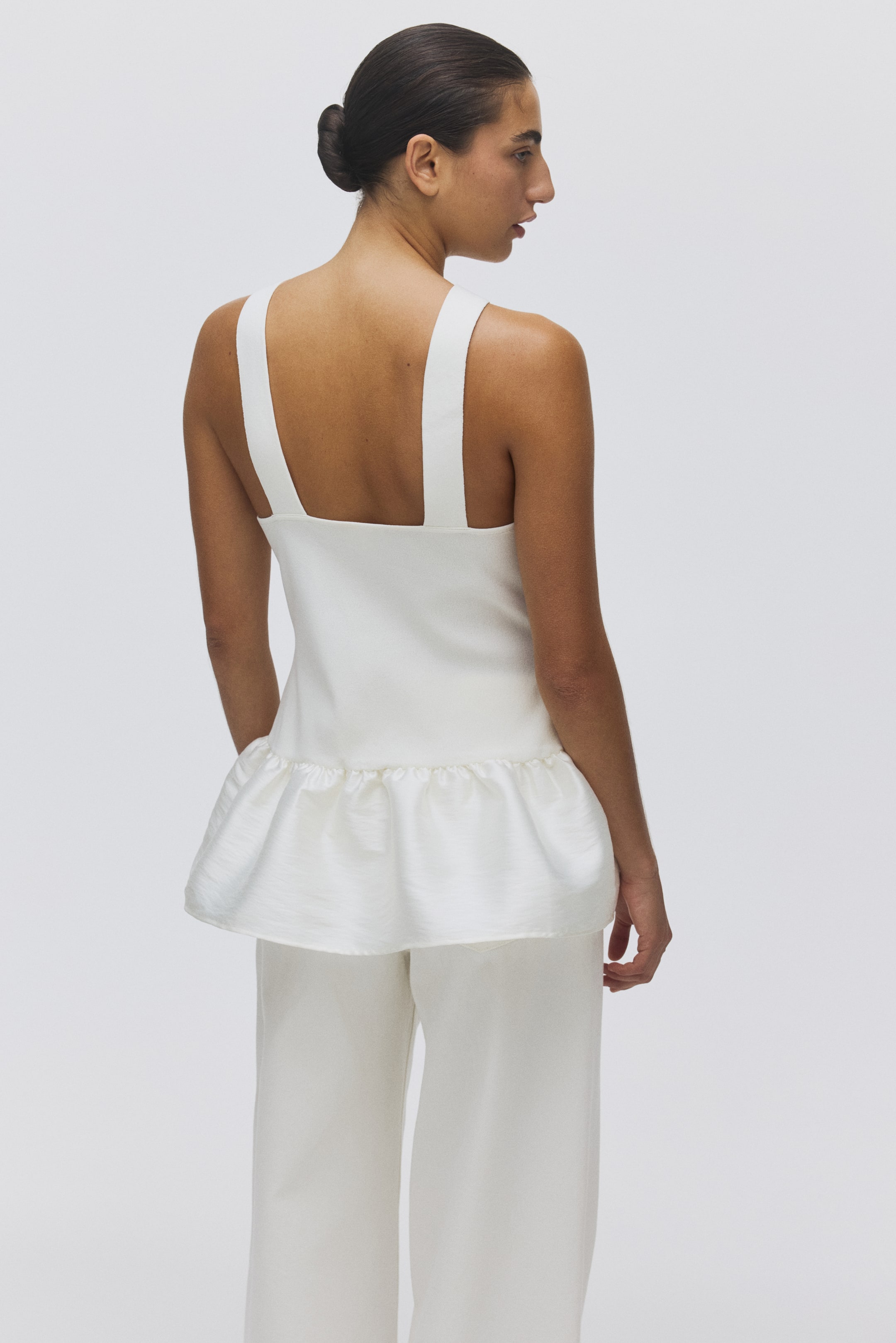View larger image: A white sleeveless peplum top and matching wide-leg pants are being presented from the back-right side. The top is designed with wide shoulder straps and a gathered, textured peplum hem, while the pants feature a relaxed, wide-leg fit.