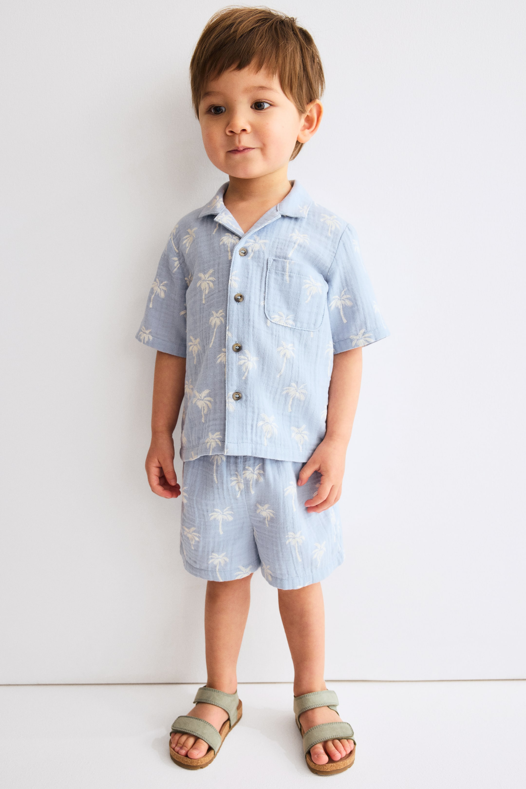 View larger image: A light blue woven fabric shirt and shorts set, featuring a light-coloured palm tree pattern, is presented from the front. The straight-cut shirt displays a resort collar, buttons down the front, short sleeves, and an open chest pocket, while the matching shorts show soft, adjustable elastication at the waist.