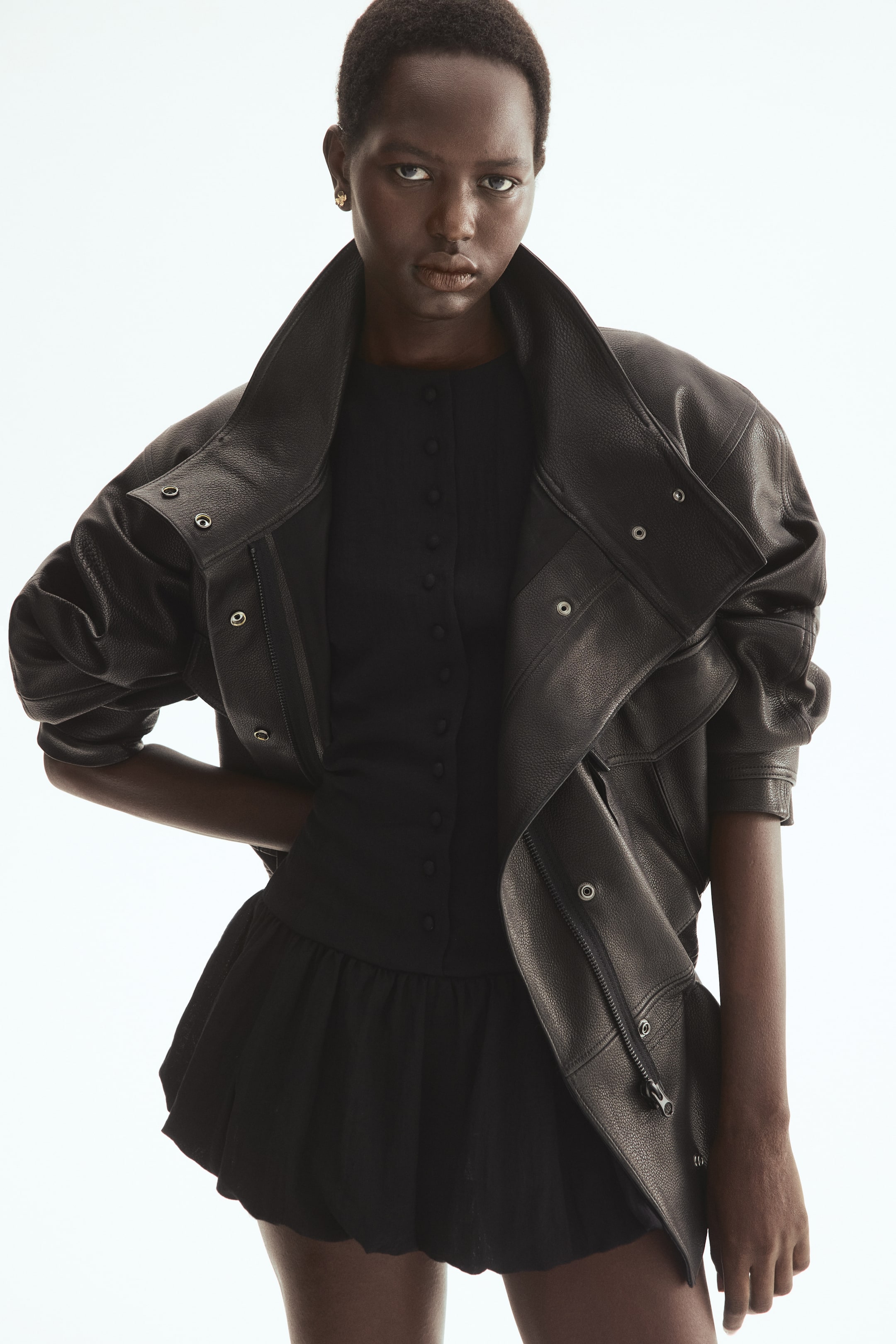 הצגת תמונה גדולה יותר: שמלה קצרה שחורה עם מחשוף עגול וכפתורים מכוסים לאורך החזית נלבשת על ידי הדוגמנית. השמלה כוללת שרוולים קצרים נפוחים ושולי בועה מקובצים, הנראים מתחת למעיל עור שחור שהדוגמנית לובשת מעל השמלה.