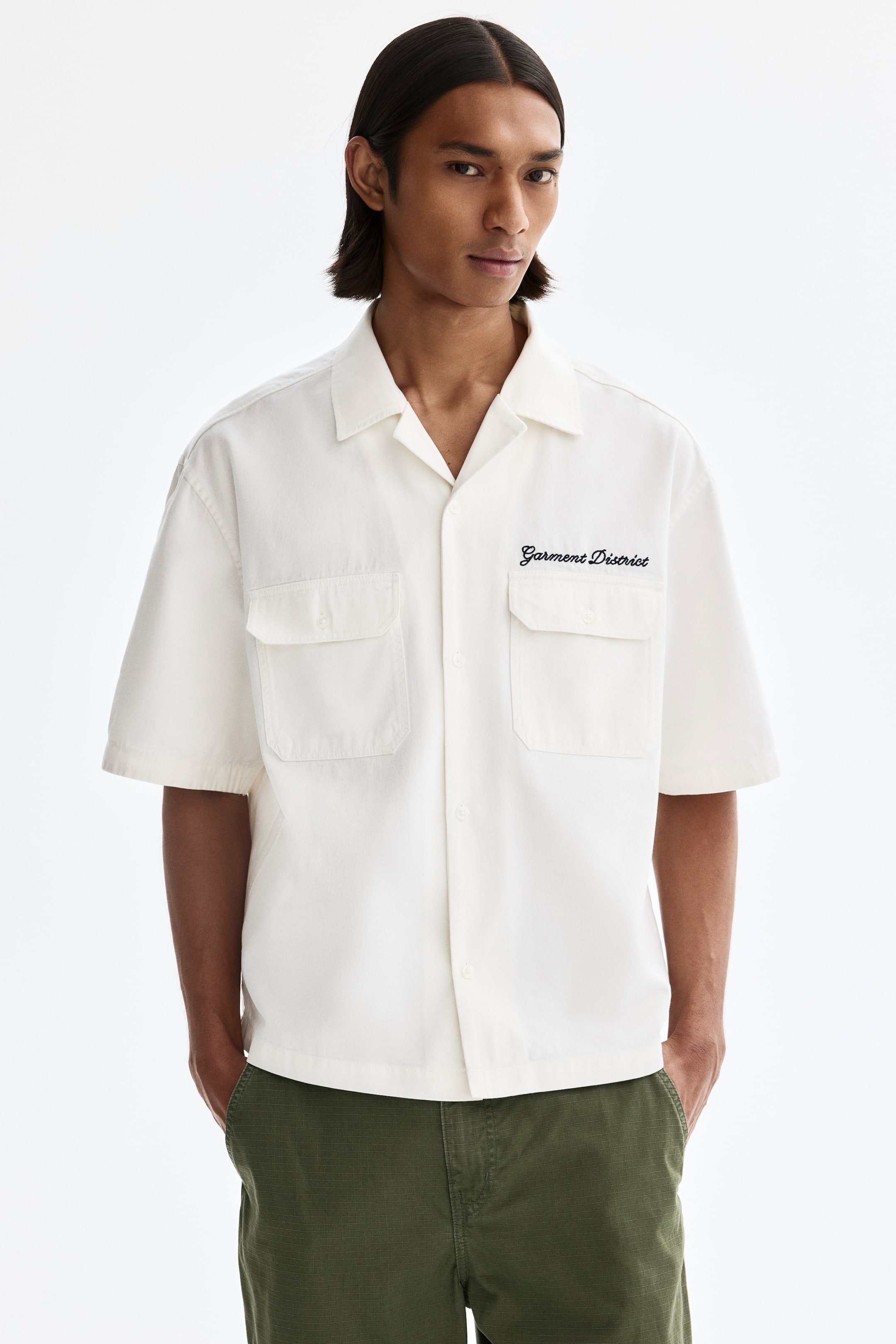 View larger image: An off-white short-sleeved shirt features a revere collar and a button-front closure, shown facing forward. It displays two chest patch pockets with flap closures and black embroidered text "Garment District" on the right chest.