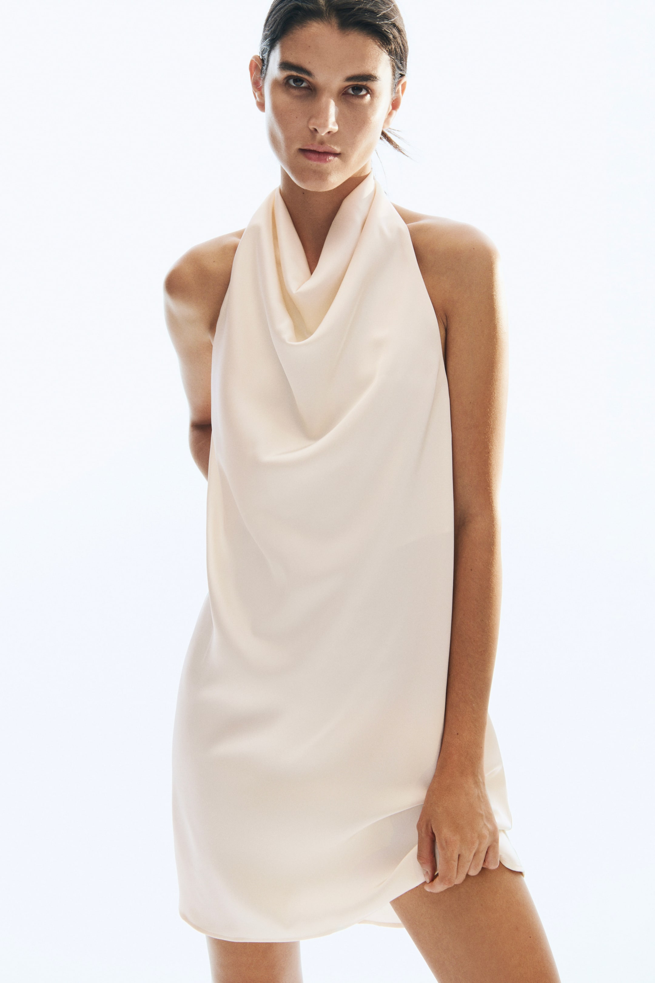 View larger image: A cream-coloured, loose-fit satin mini dress featuring a draped cowl neckline and halter neck is being worn by the model. The model is holding the lower hem of the dress with one hand.