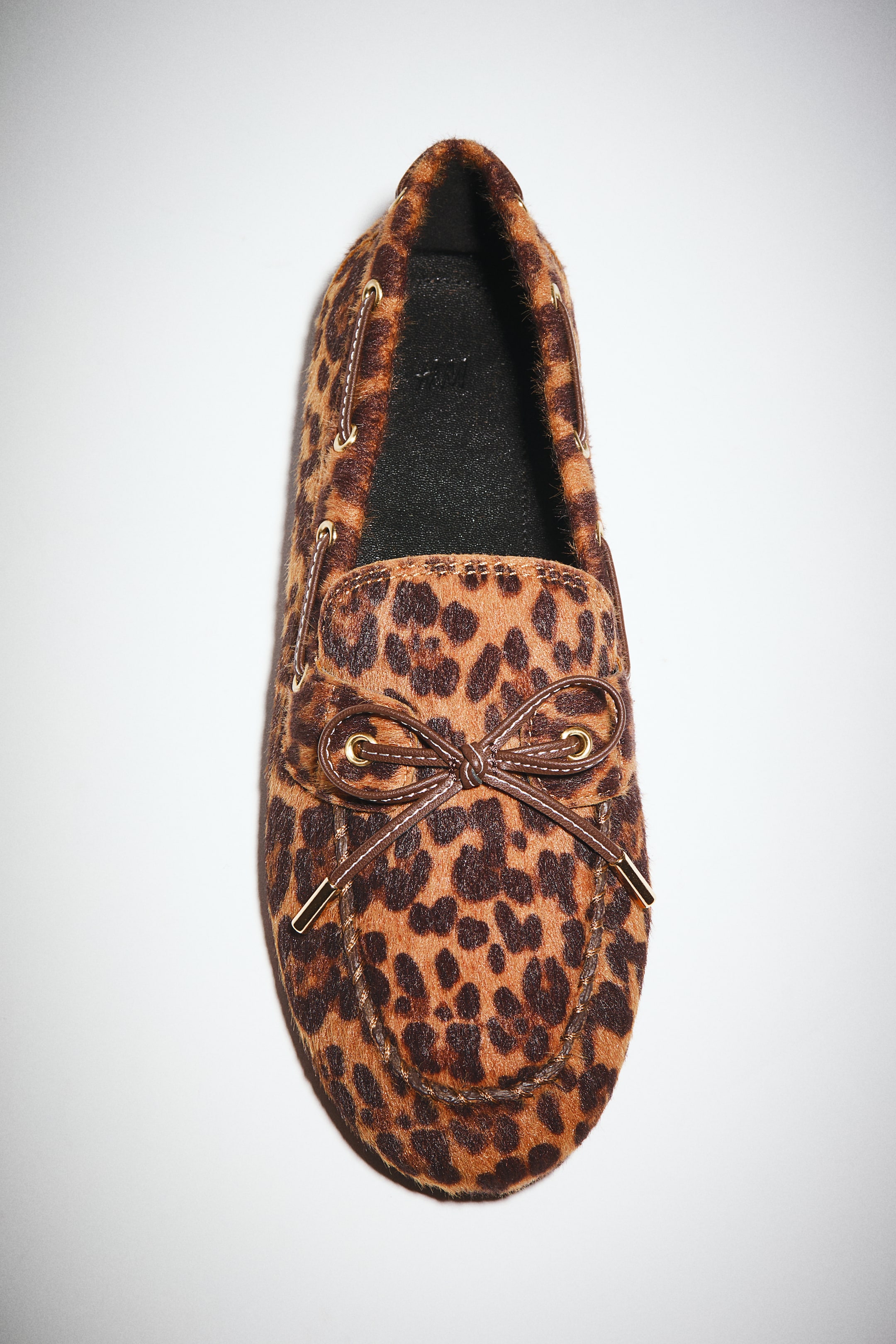 View larger image: A leopard print driving moccasin is facing forward, featuring brown and black spots on a tan textured material. Brown leather laces are threading through gold eyelets along the sides and tying into a bow on the vamp, with gold aglets visible on the lace ends.
