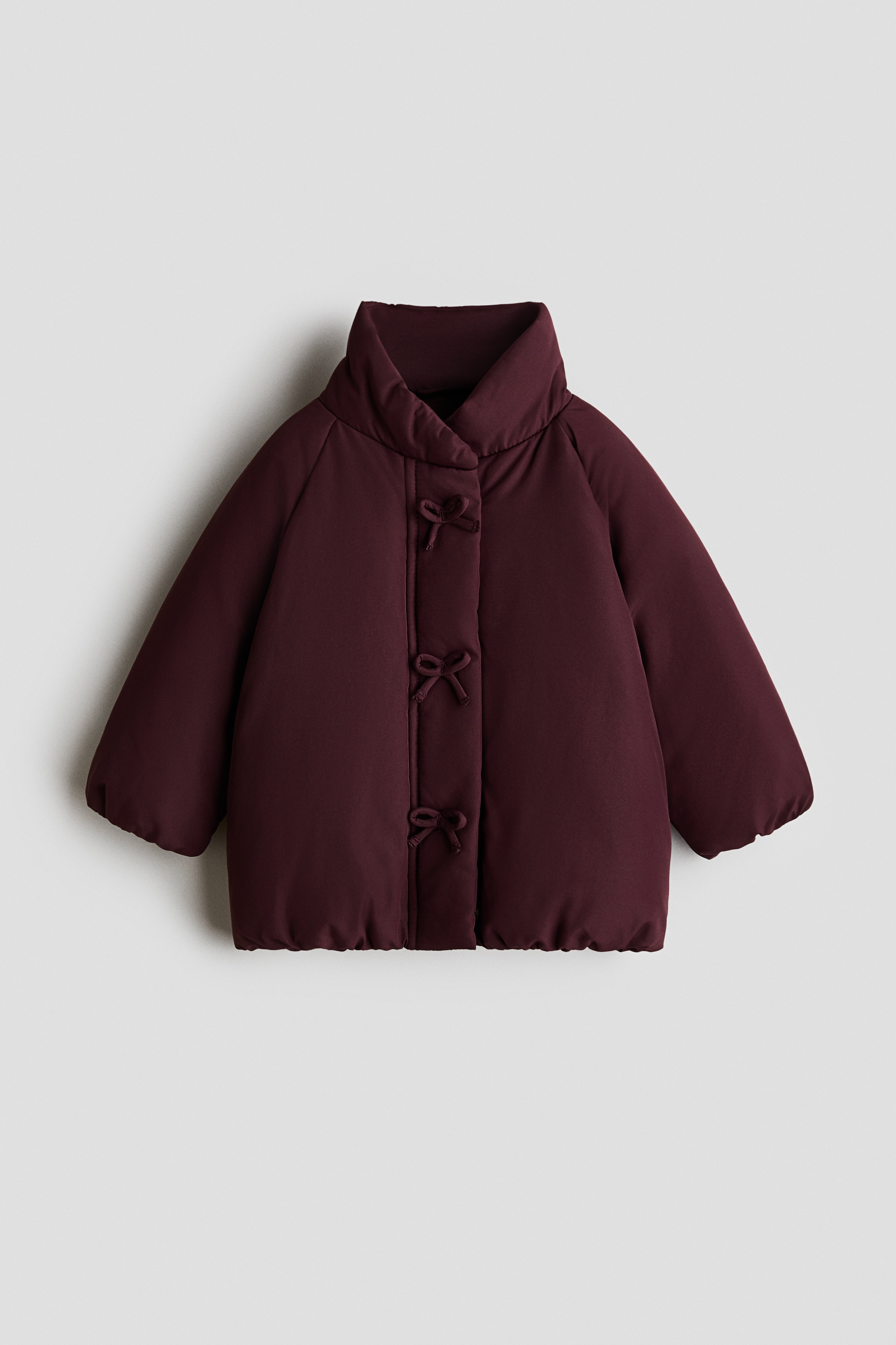 A dark burgundy quilted jacket, shown from the front, featuring a high stand-up collar, raglan sleeves, and three decorative fabric bows aligned vertically down the front wind flap.
