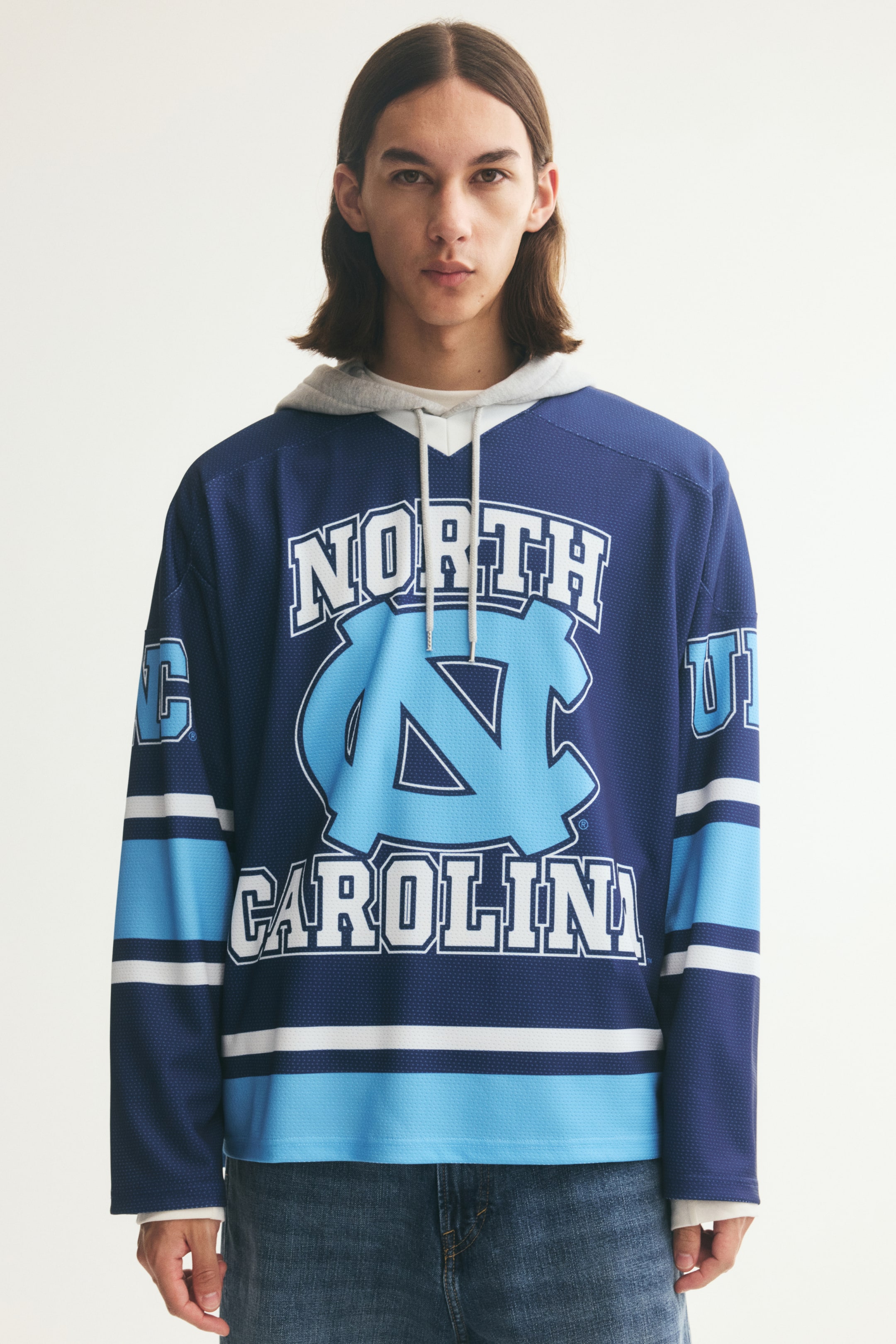 Vezi imaginea mai mare: O cămașă oversized cu mânecă lungă, în stil hockey, din plasă bleumarin închis, este purtată de model peste un hanorac gri deschis. Cămașa are un grafic „North Carolina” în albastru deschis și alb pe piept, un decolteu în V cu striații și umeri căzuți.