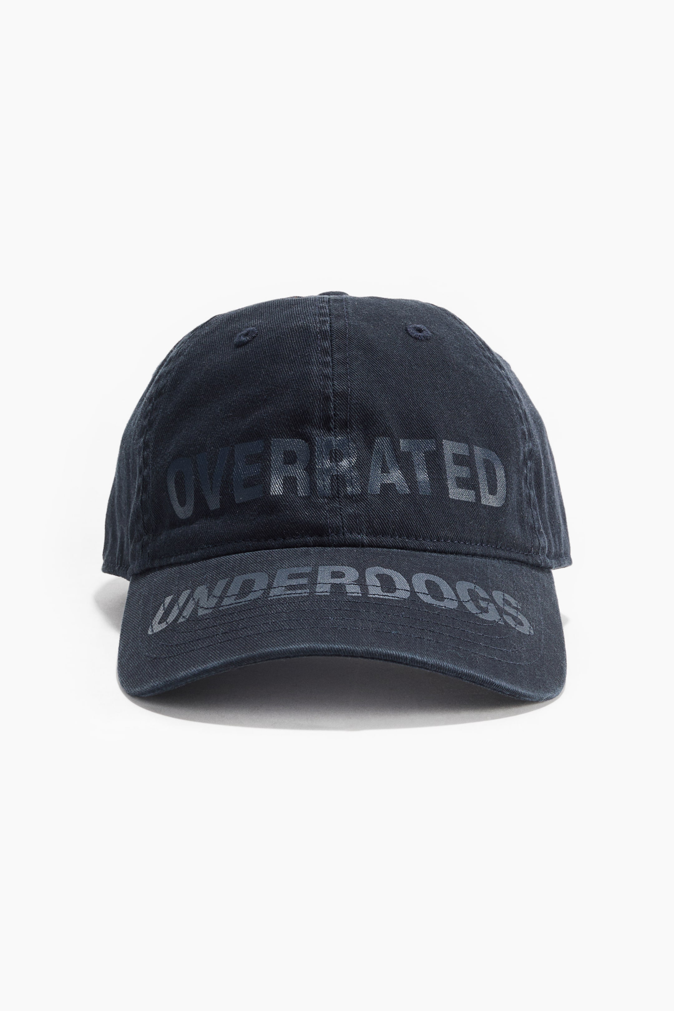 View larger image: A dark blue cotton twill cap is facing forward, displaying "OVERRATED" on its crown and "UNDERDOGS" on its curved brim. Both phrases appear in a subtly darker, glossy print, and two eyelets are visible on the cap's front panels.