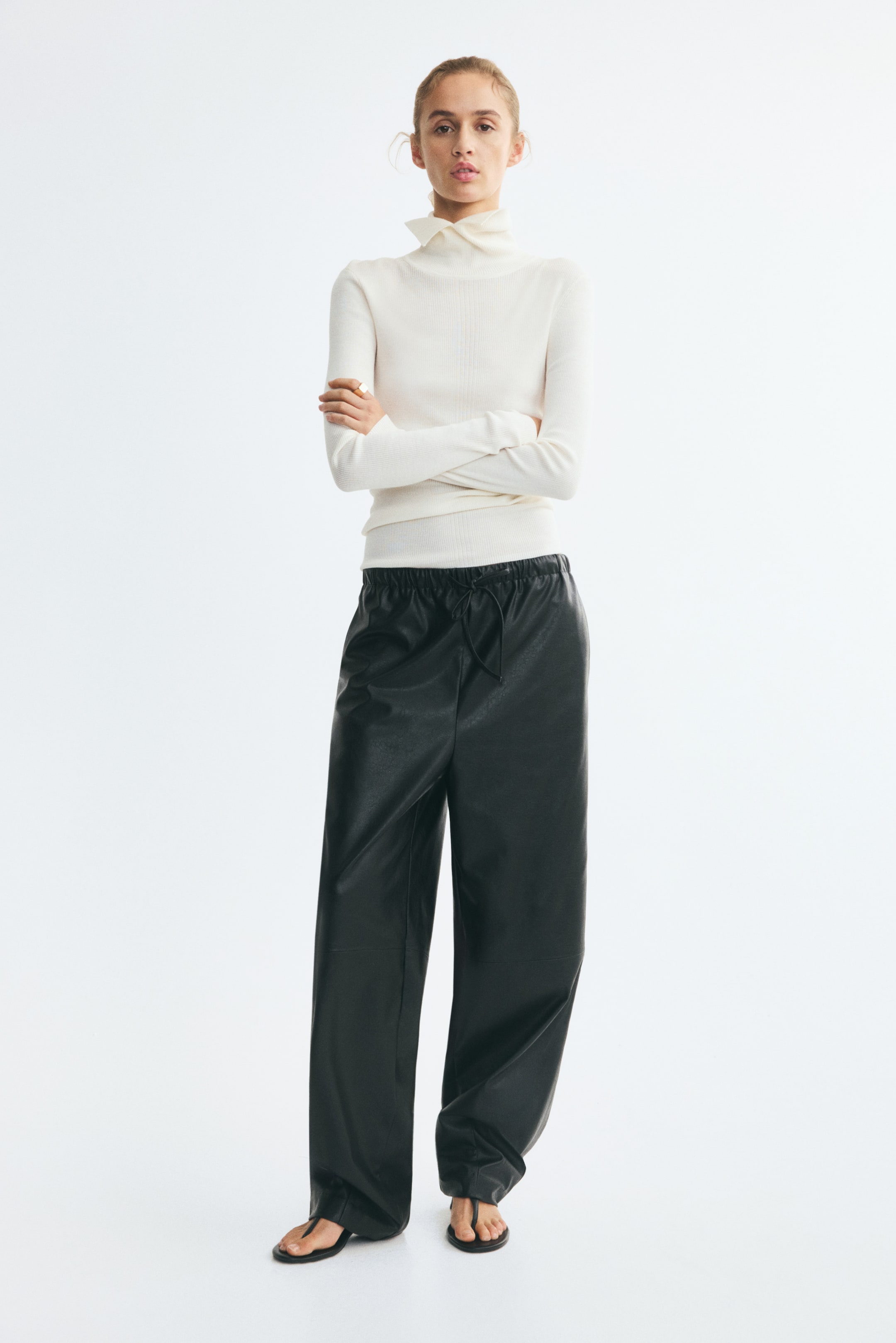 Agrandir l'image: Le pantalon ample en tissu enduit noir, doté d'une taille haute élastiquée avec cordon de serrage et de jambes larges de forme barrel, est porté par le mannequin. Le pantalon est associé à un haut crème à col roulé et manches longues, ainsi qu'à des sandales plates noires.