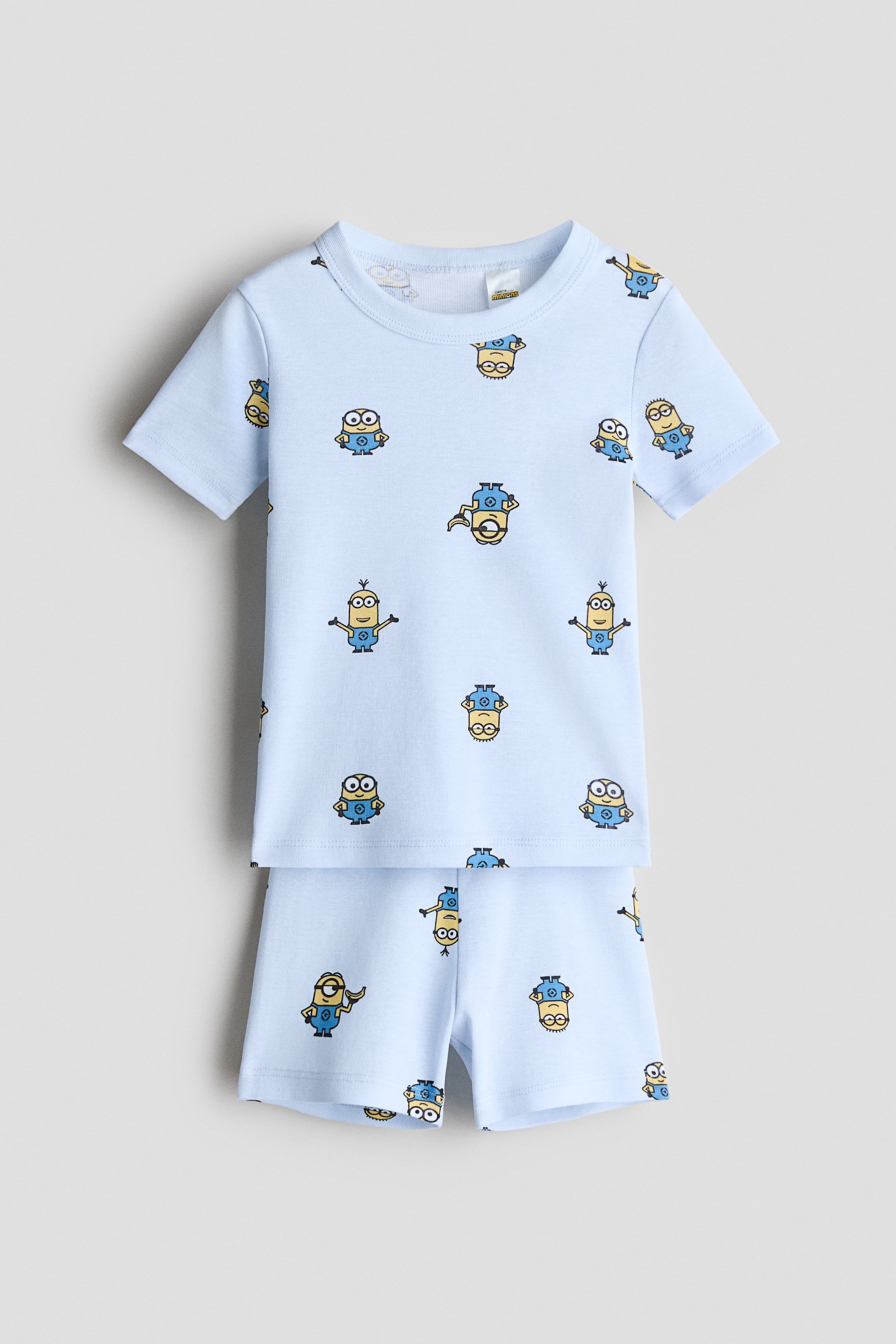View larger image: A pajama set, shown from the front, features a light blue T-shirt and matching shorts, both crafted from soft cotton jersey. The short-sleeved T-shirt and shorts showcase an all-over print of yellow Minions characters, with the shorts also having an elastic waistband.