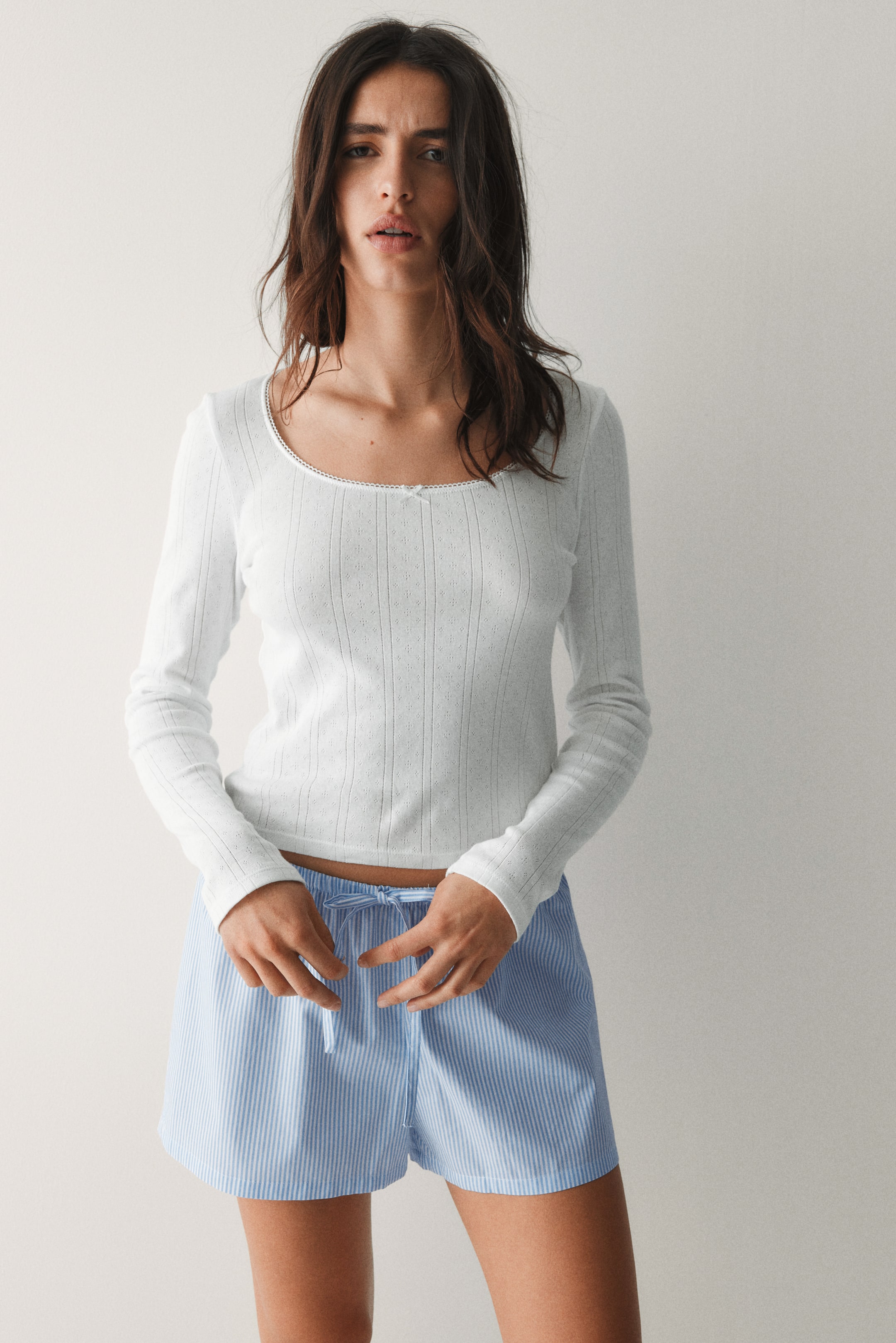 Xem ảnh lớn hơn: Áo dài tay màu trắng có cổ tròn với viền ren và một chiếc nơ nhỏ, thể hiện hoa văn tinh tế với các đường kẻ dọc và các lỗ nhỏ. Quần short kẻ sọc xanh và trắng được mặc, có dây rút màu trắng ở eo.