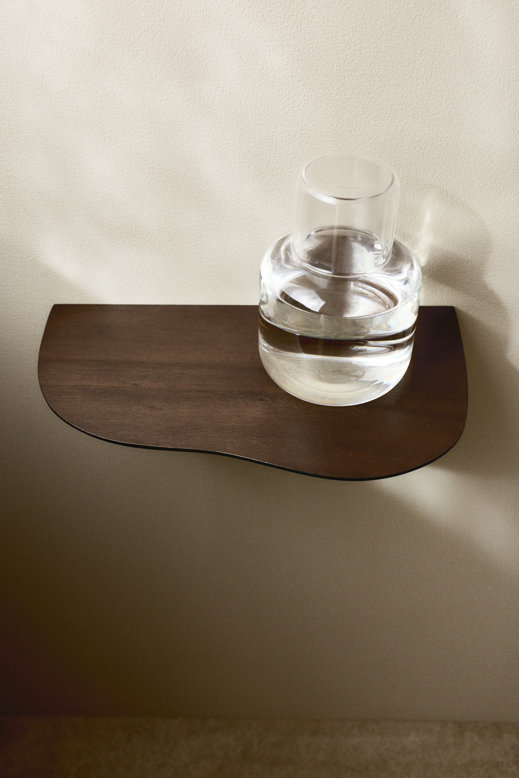 Ver imagen más grande: Una estantería de madera marrón oscuro está montada horizontalmente, mostrando su borde frontal suavemente curvado en forma de S y sus extremos redondeados. Un decantador de vidrio transparente con una taza de vidrio a juego en la parte superior descansa en su lado derecho.