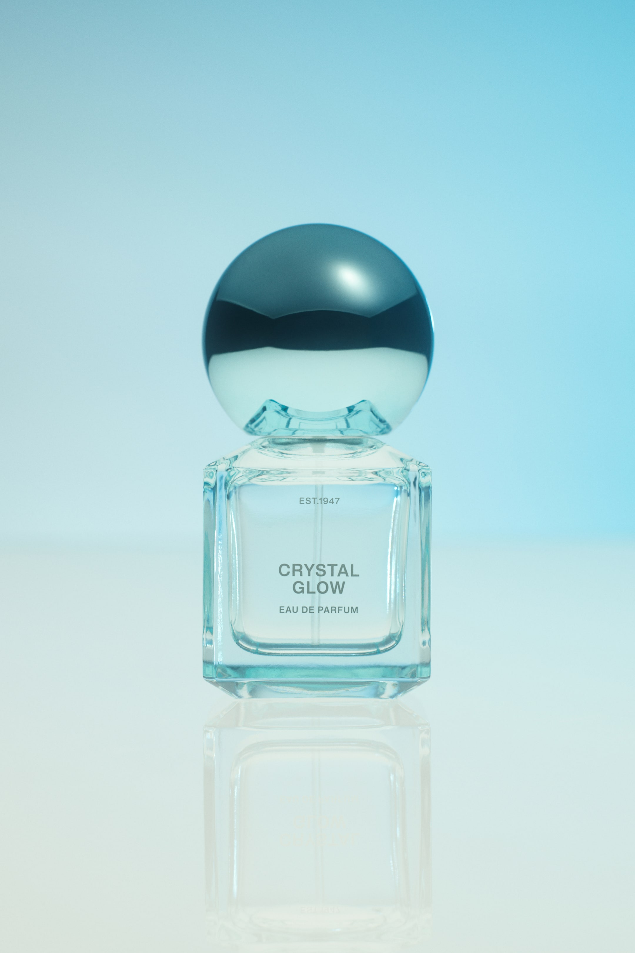 View larger image: A front-facing Crystal Glow Eau de Parfum bottle is standing upright, featuring a clear, square glass body with bevelled corners and a large, reflective blue spherical cap. The front of the bottle displays the text "EST.1947," "CRYSTAL GLOW," and "EAU DE PARFUM" in a clean grey font. The bottle rests on a glossy surface that reflects its design.