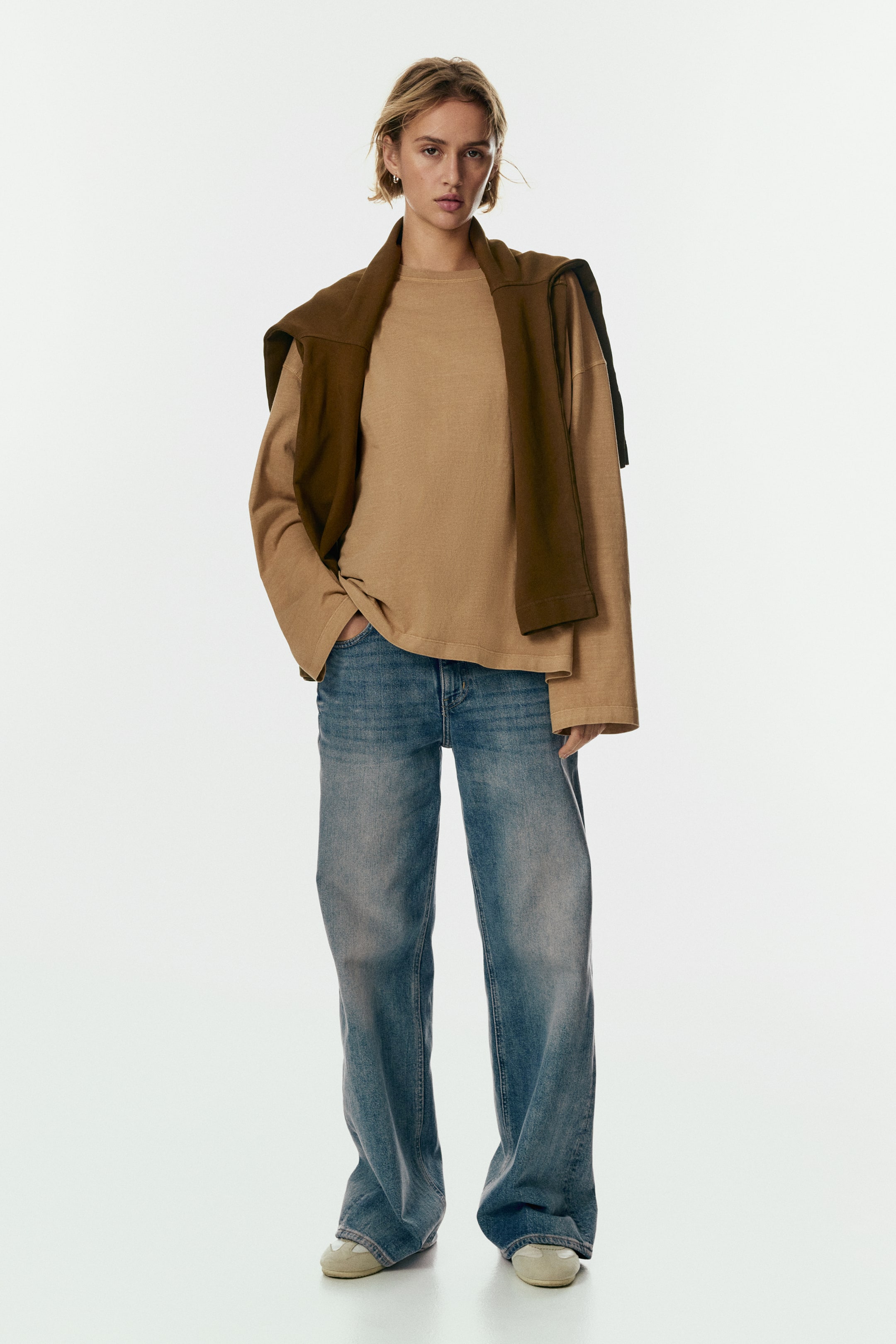 Xem ảnh lớn hơn: Một chiếc áo phông dài tay màu nâu nhạt, cổ tròn, có dáng rộng rãi đang được mặc cùng với quần jean ống rộng, màu nhạt, hơi đọng lại ở mắt cá chân. Một chiếc áo len màu nâu ô liu đậm được khoác hờ qua vai.