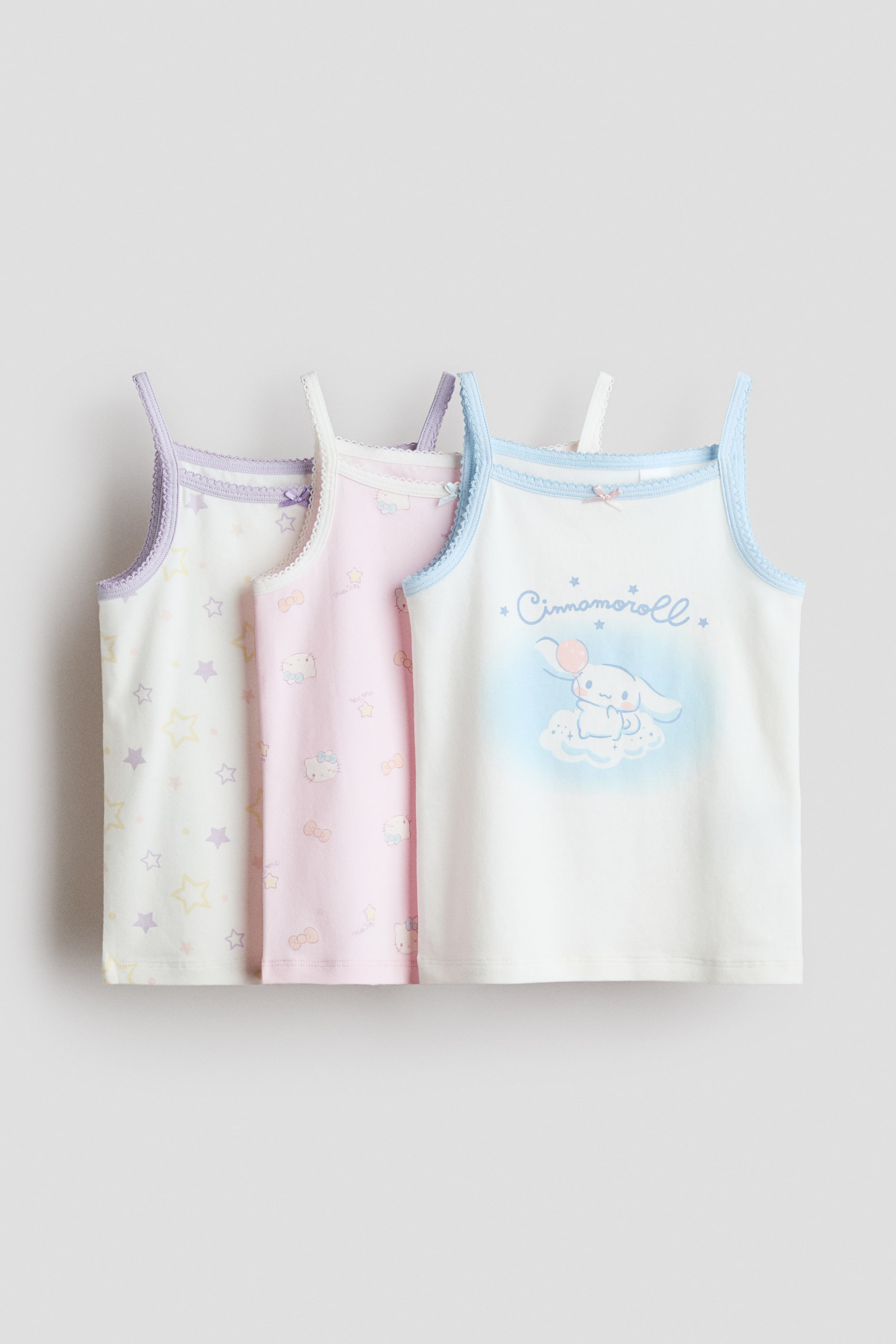 View larger image: A set of three camisole tops is displayed with their front sides facing forward, arranged side-by-side. The top on the left is white with pastel stars and purple trim. In the centre, the top is pink featuring Hello Kitty patterns with white trim. On the right, the top is white showcasing a Cinnamoroll graphic with blue trim. Each top is designed with thin straps, scalloped edges along the neckline and armholes, and a small bow at the centre front.