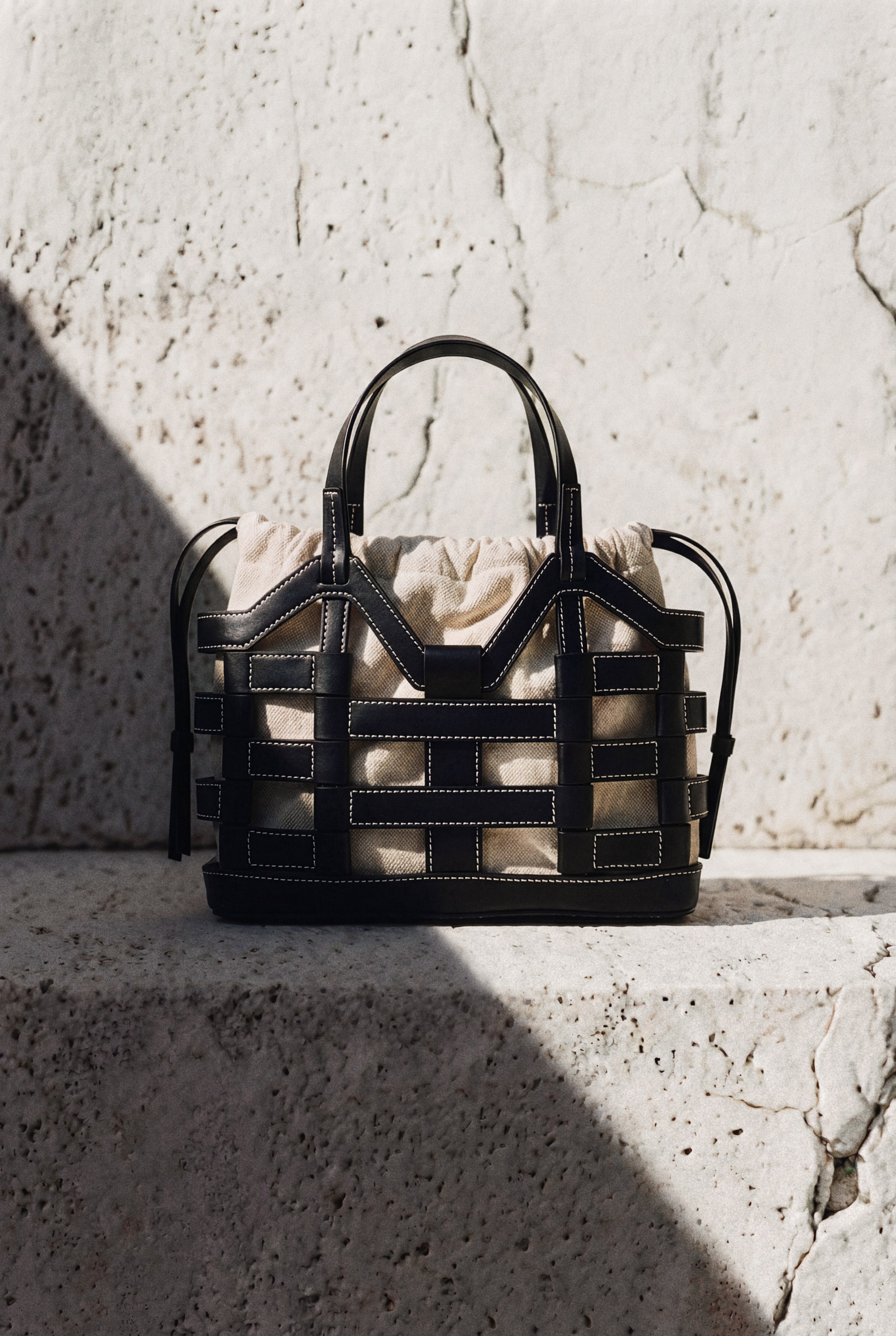 View larger image: Presenting a black woven leather tote bag showcasing its front, accentuated by white contrast stitching that highlights the straps, forming an open-weave design. Inside, a light-colored gathered fabric drawstring pouch is visible, while two black top handles extend upwards.