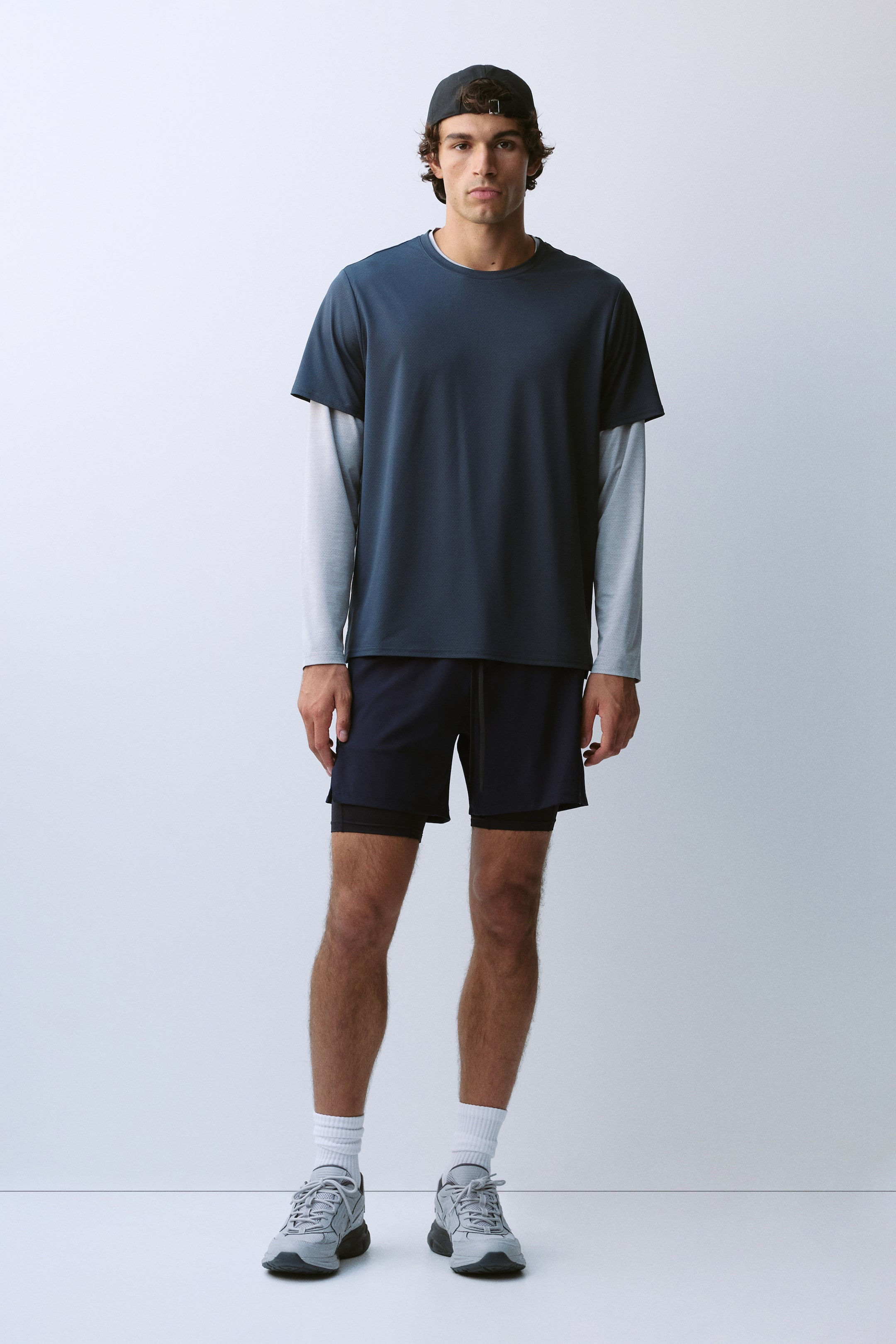 Ver imagen más grande: La modelo lleva una camiseta deportiva de manga corta en verde azulado oscuro, superpuesta sobre una prenda de manga larga en gris claro. El conjunto se completa con pantalones cortos deportivos en azul marino oscuro con forro interior negro, calcetines blancos de caña media y zapatillas grises.