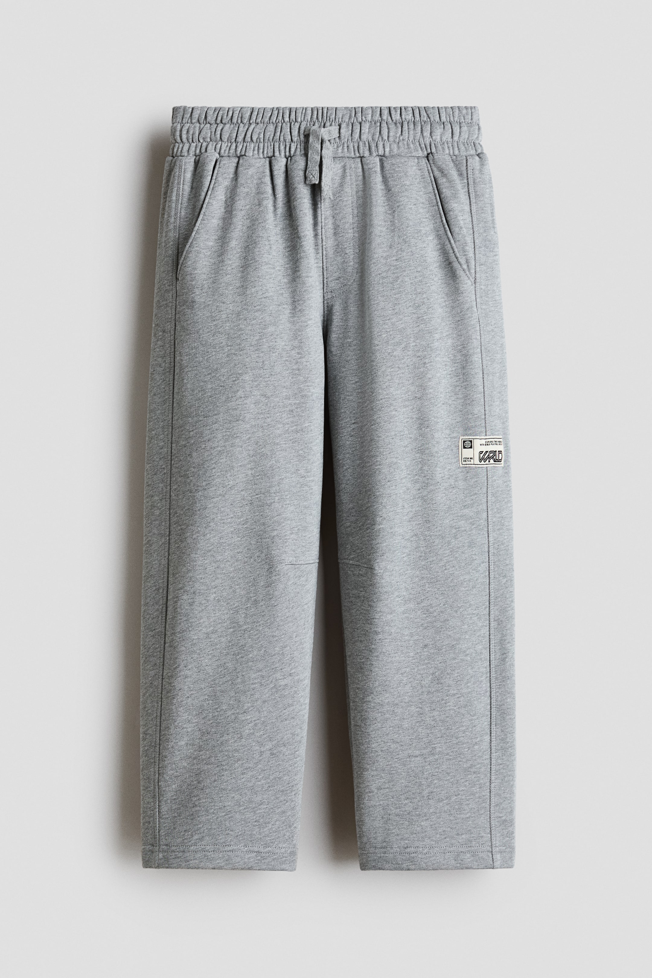 View larger image: Grey sweatpants are displayed from the front, featuring an elasticated waistband with a matching drawstring and side slip pockets. The straight-leg design shows vertical seam detailing down the sides, horizontal knee seams, and a small rectangular graphic patch on the lower left leg.