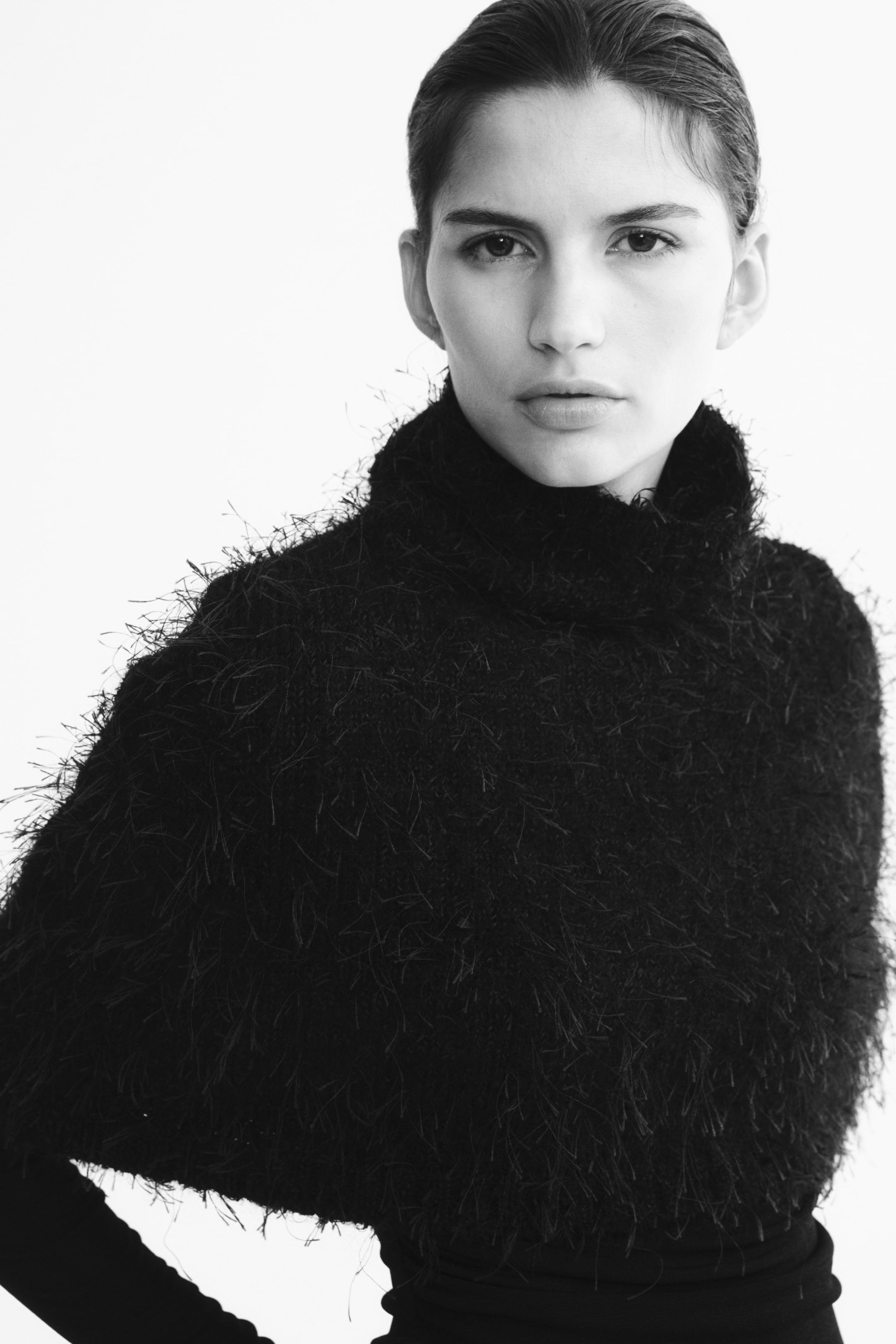 Agrandir l'image: Une cape courte et oversize en maille noire texturée et duveteuse est portée par le mannequin par-dessus un haut ajusté à manches longues. La cape présente un col montant et un ourlet droit, drapée souplement sur les épaules.